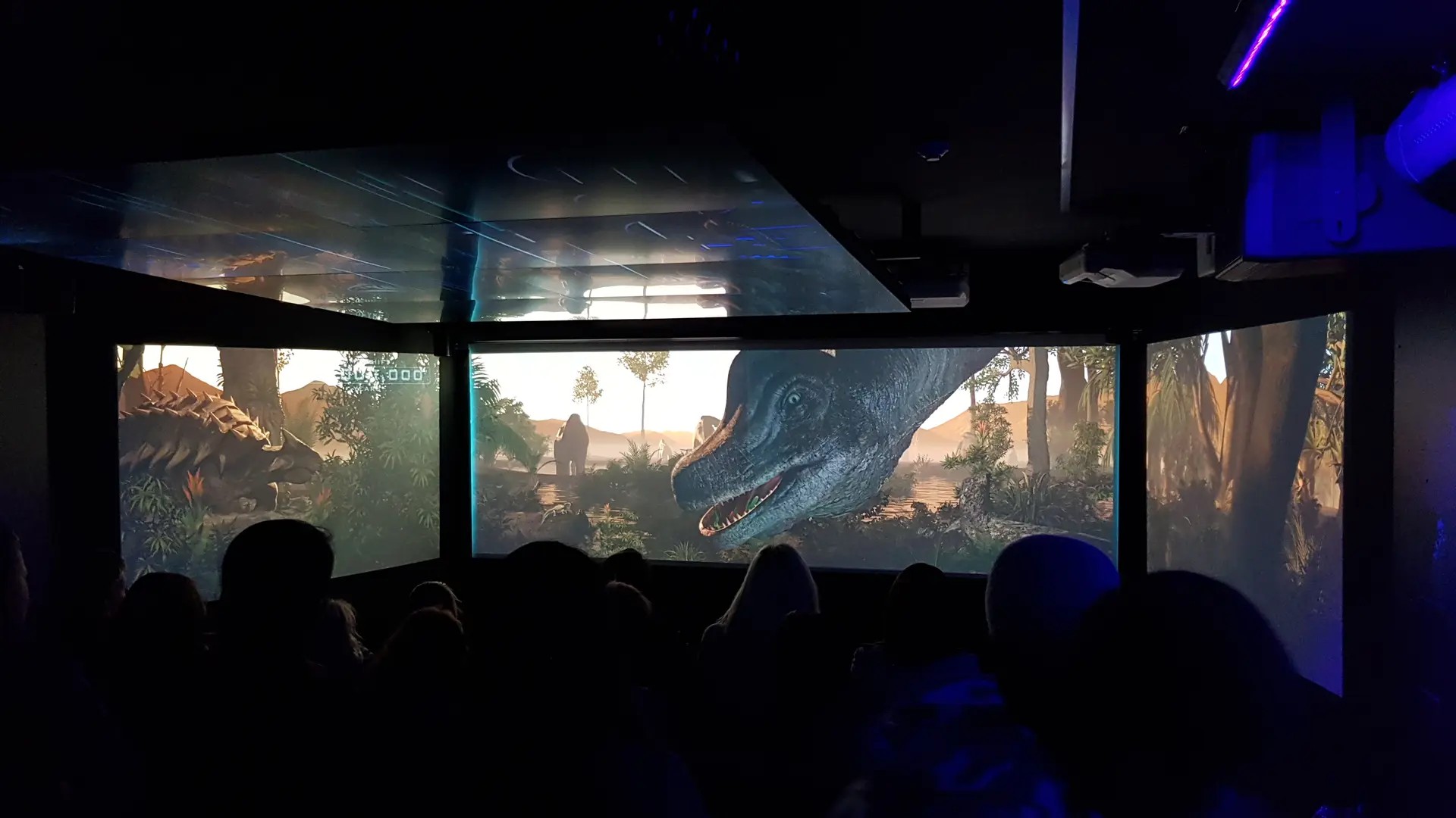 Dark cinema space underground with three continuous screens. A dinosaur is peeking through the middle screen.