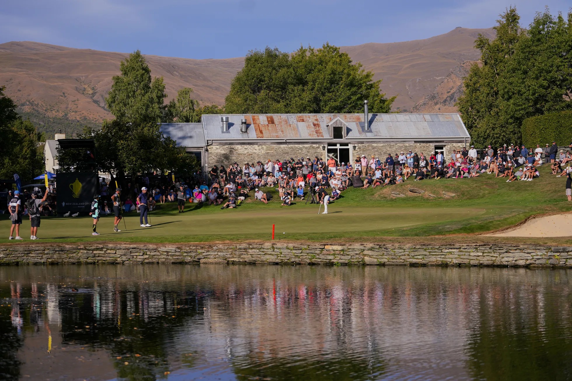 Crowd of people watch a golf game at a resort by a lake.
