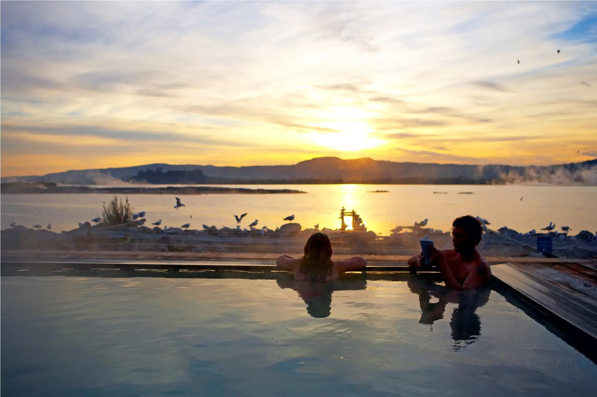 Couple relaxing in a private Polynesian spa pool by the lakefront at sunset.