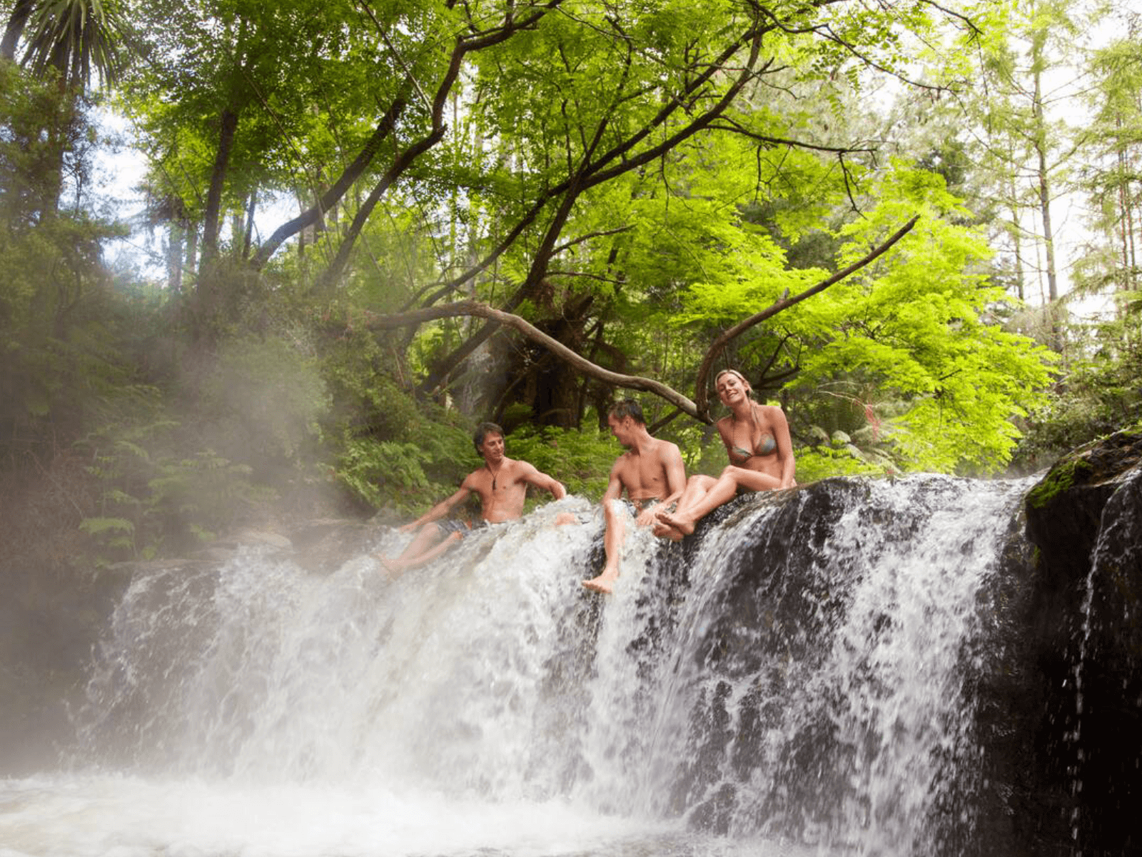 Three friends relaxing on a natural hot spring waterfall in a lush Rotorua forest, surrounded by steam and native greenery.