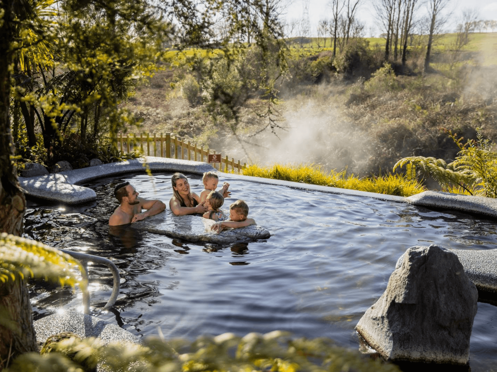 Family relaxing in a natural geothermal hot pool at Waikite Valley, surrounded by steam, native plants and countryside views.