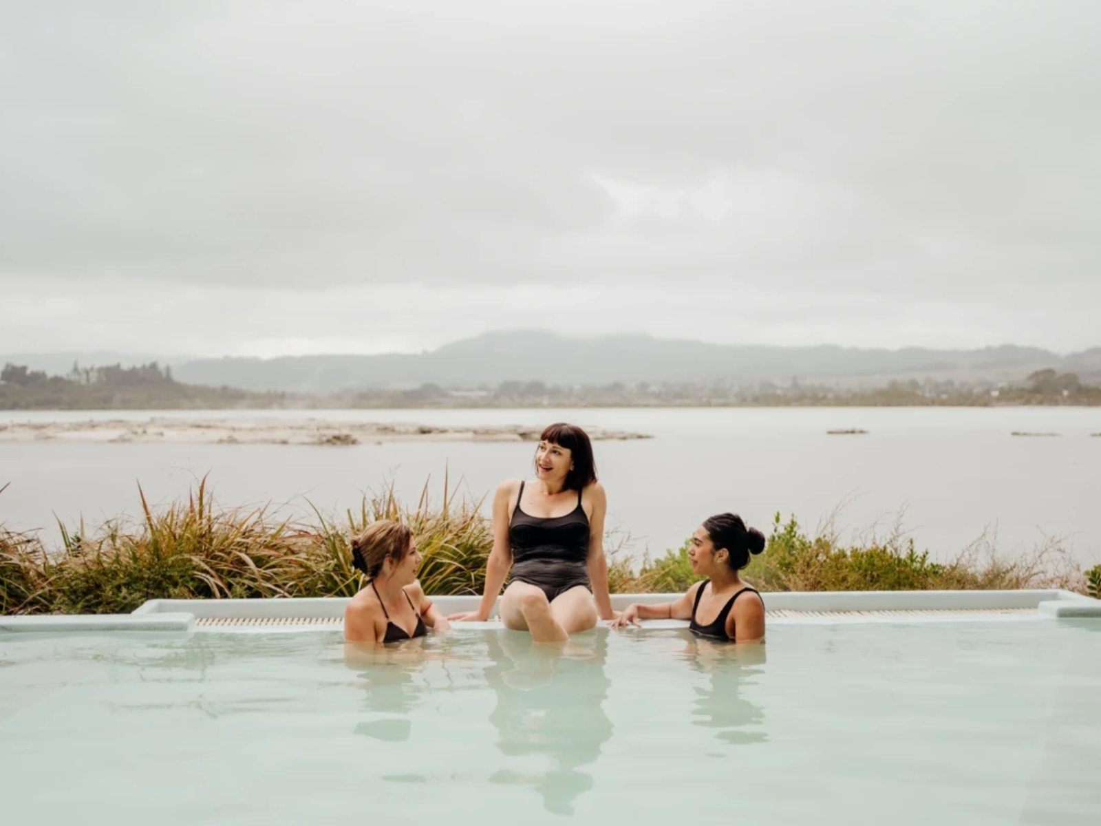 Three women relaxing in a geothermal hot pool overlooking Lake Rotorua on a cloudy day, with hills in the distance.