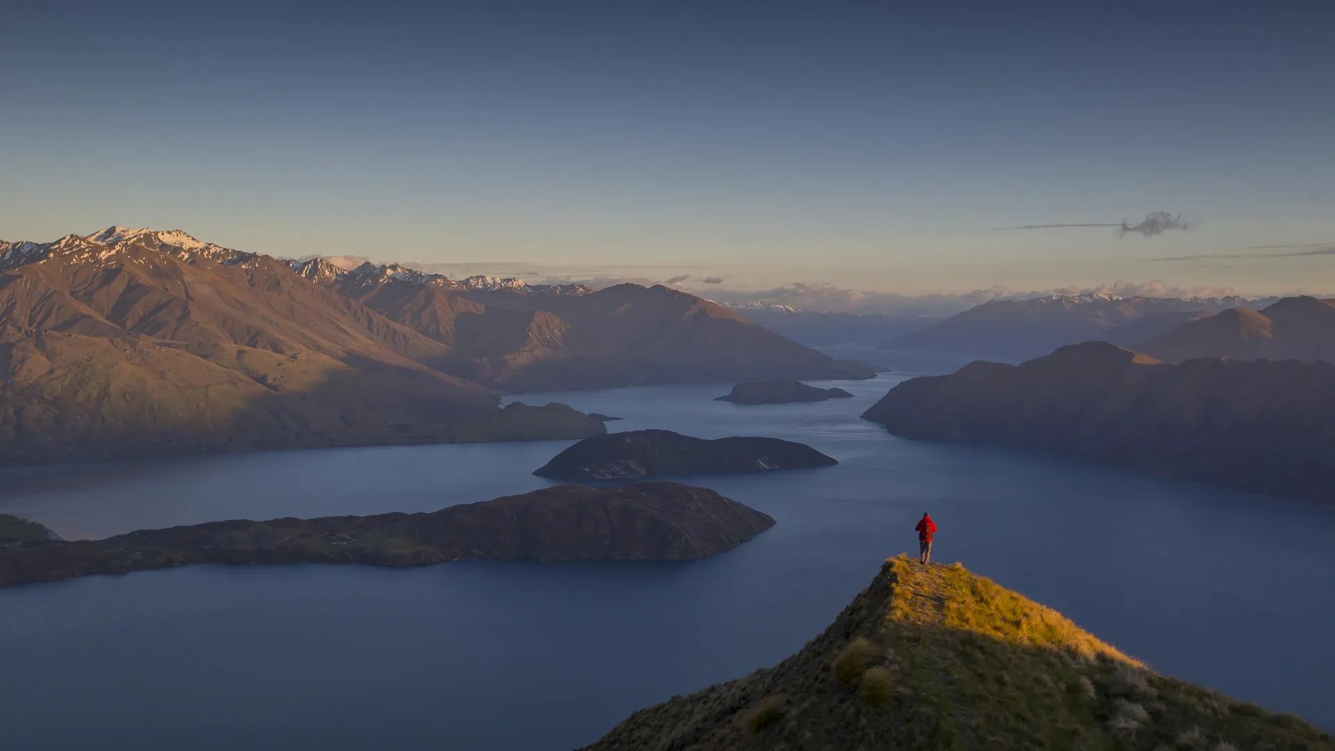 A person stands atop Roys Peak with stunning views of lake Wanaka surrounding him in every direction.