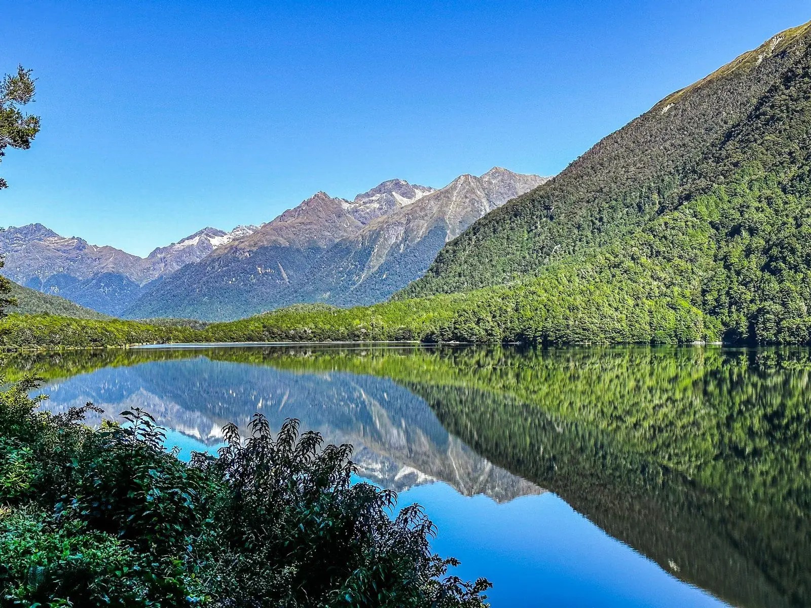Clear reflections of forested hills and rugged mountain peaks mirrored in the still waters of Lake Gunn under a bright blue sky