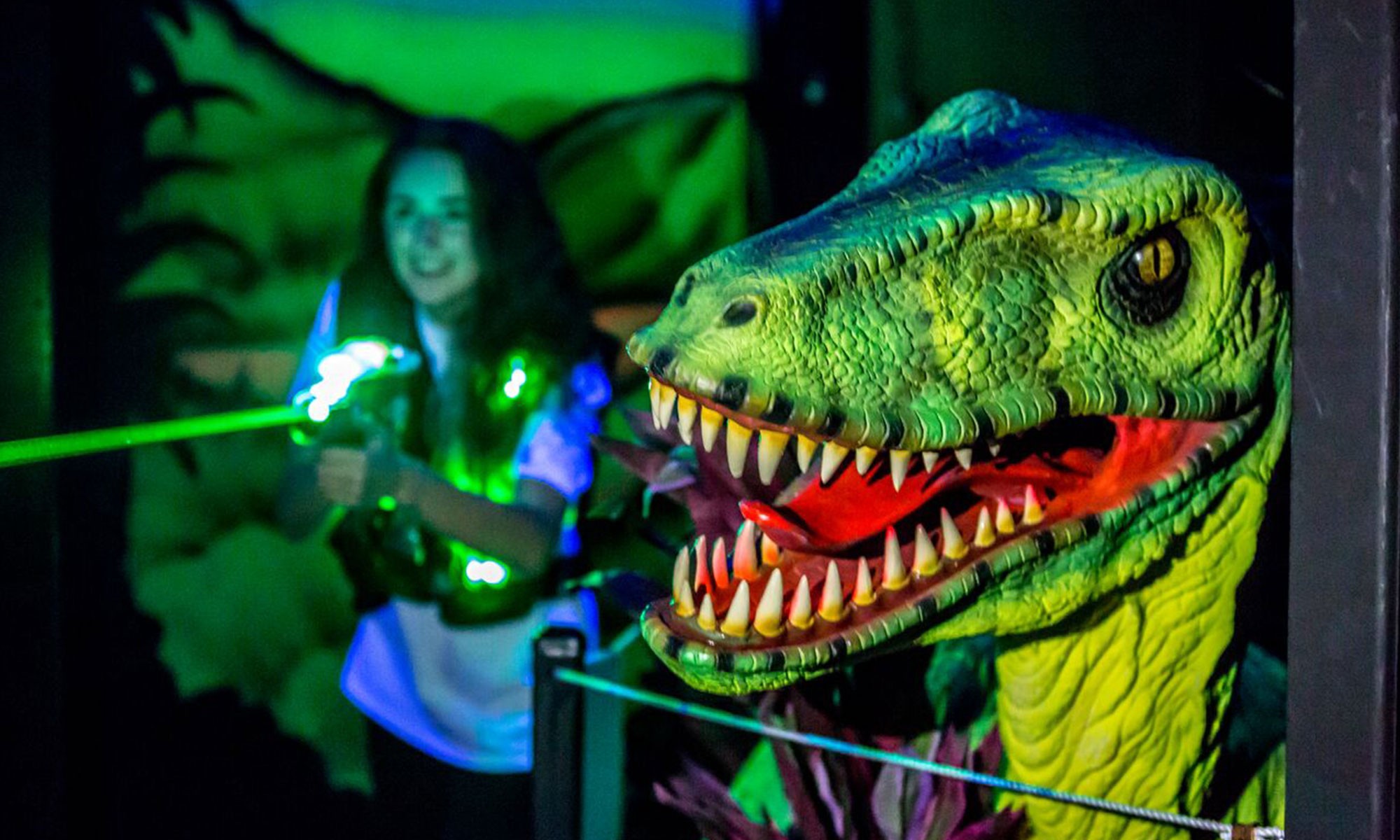 Person playing laser tag in a neon green-lit room. There's a dinosaur statue.