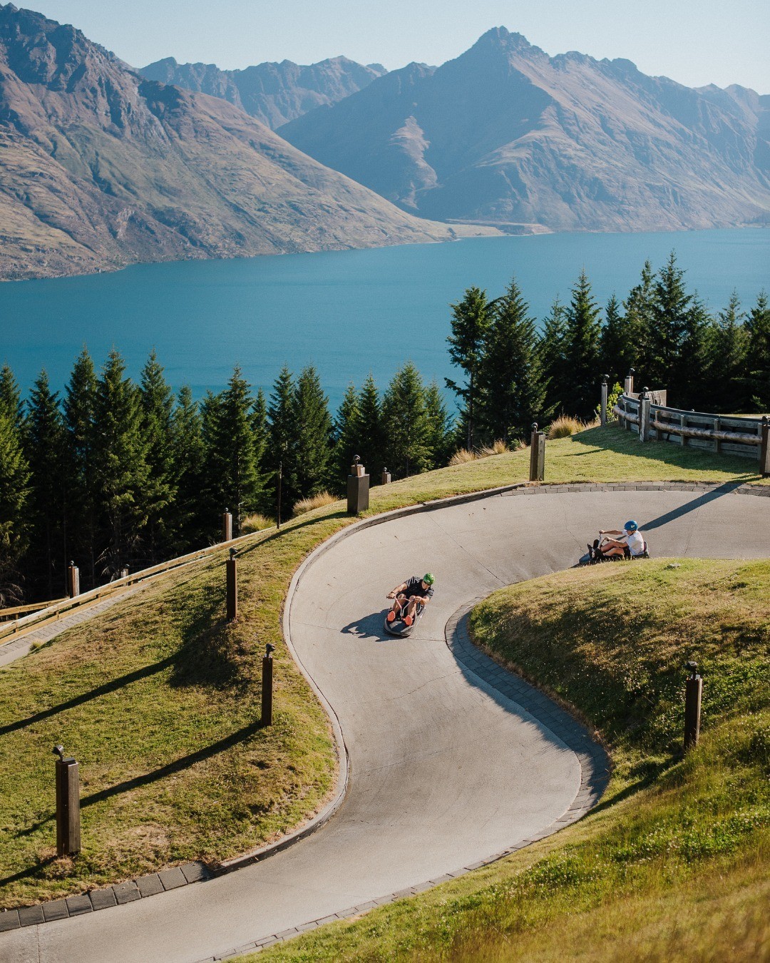 Two people ride the Skyline Queenstown Luge with stunning views of Lake Whakatipu behind them.