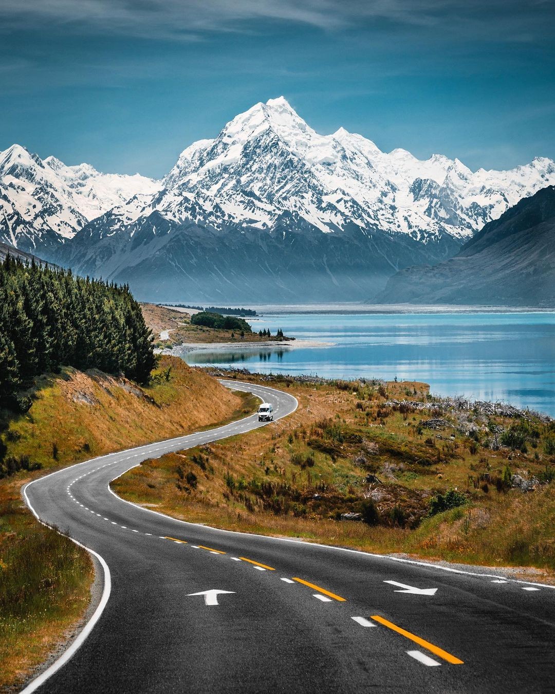 A van travels along a scenic road on the side of Lake Pukaki with mount Cook in the background.