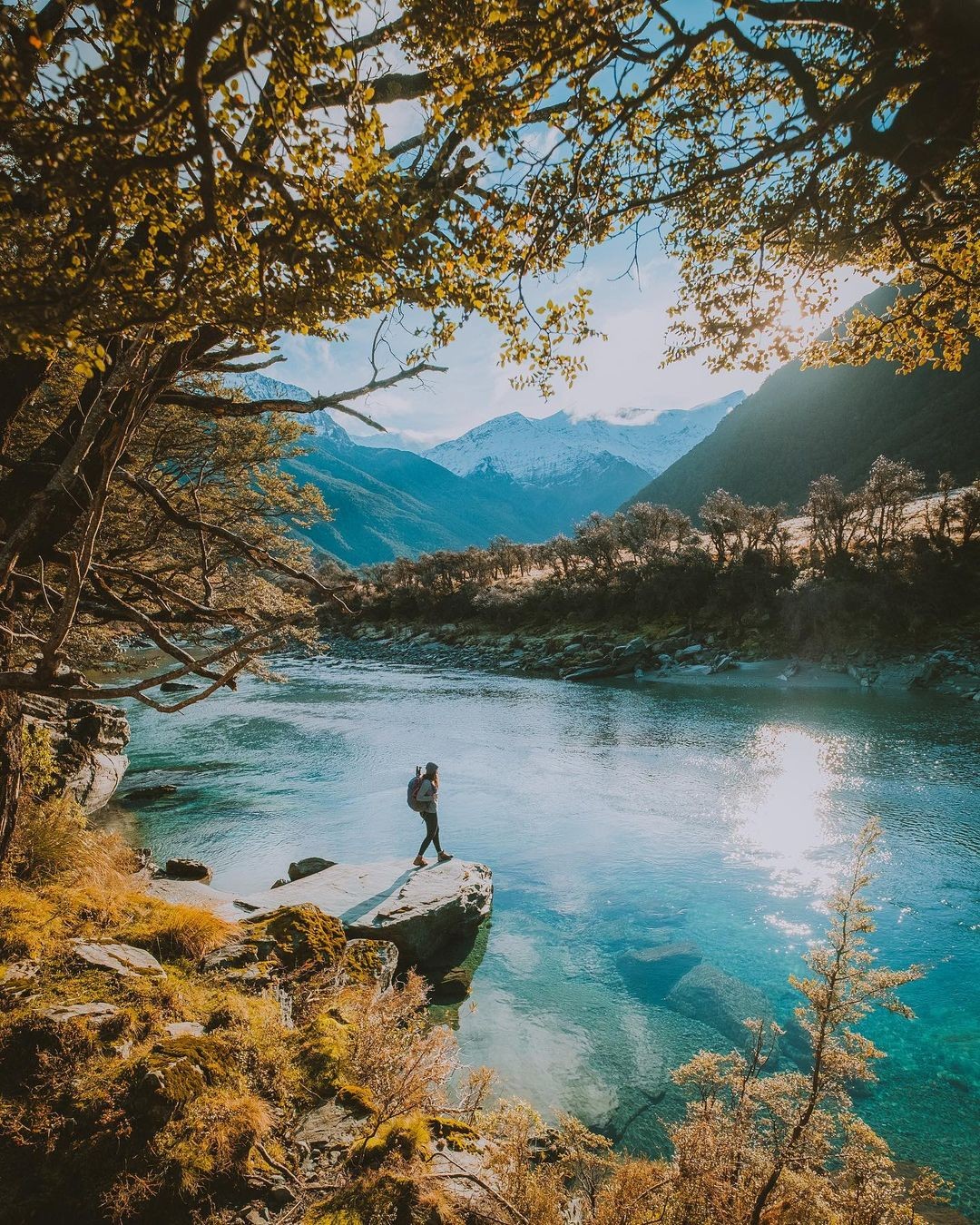 A woman stops on a large rock overhanging a pristine river flowing through a valley in the Mount Aspiring National Park.