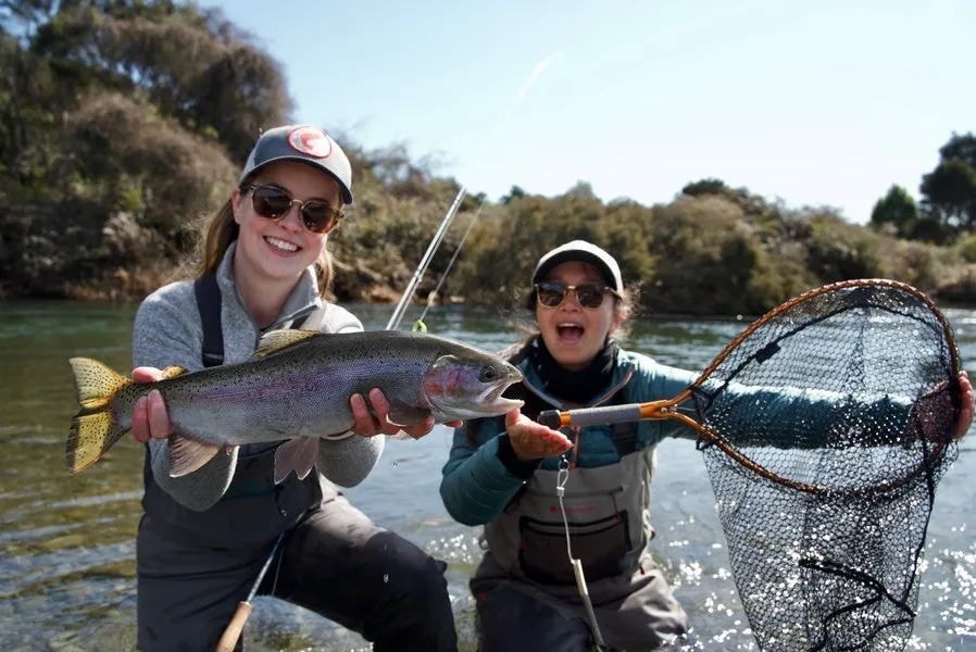 A woman shows off a large trout she has just caught whilst her friend poses next to her with a net.