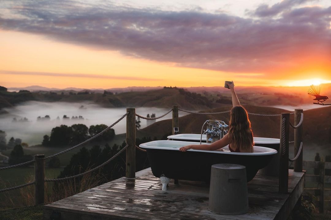 A woman raises her glass in a hot tub whilst a stunning sunset highlights the sky behind here.