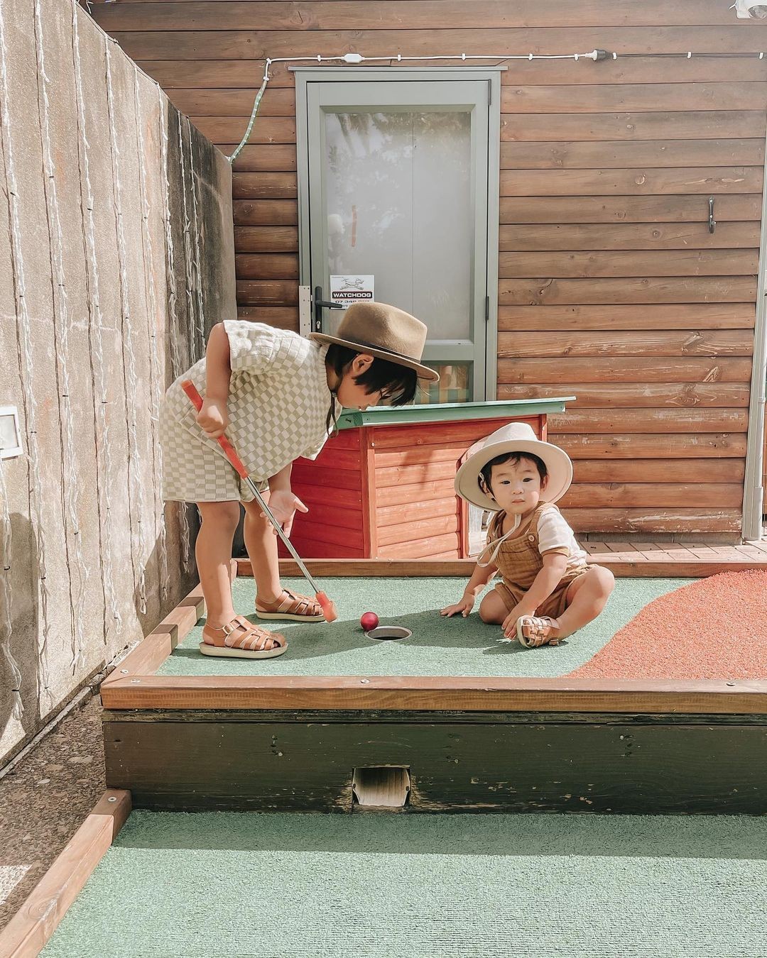 A young child tries to hit a ball into a mini putt hole whilst a baby sits on the other side.