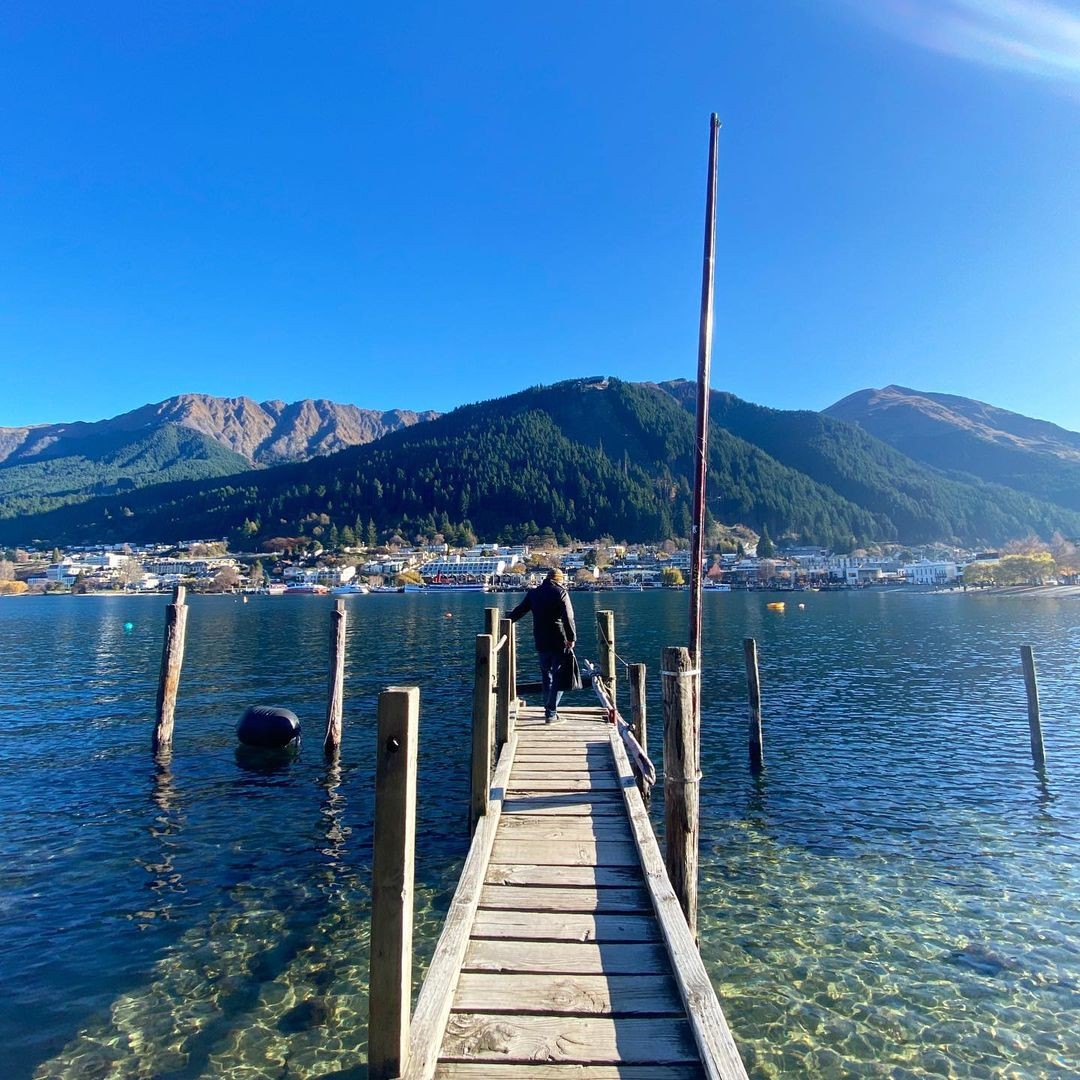 A woman stands at the end of a wharf located in the Queenstown Gardens looking back at downtown Queenstown.