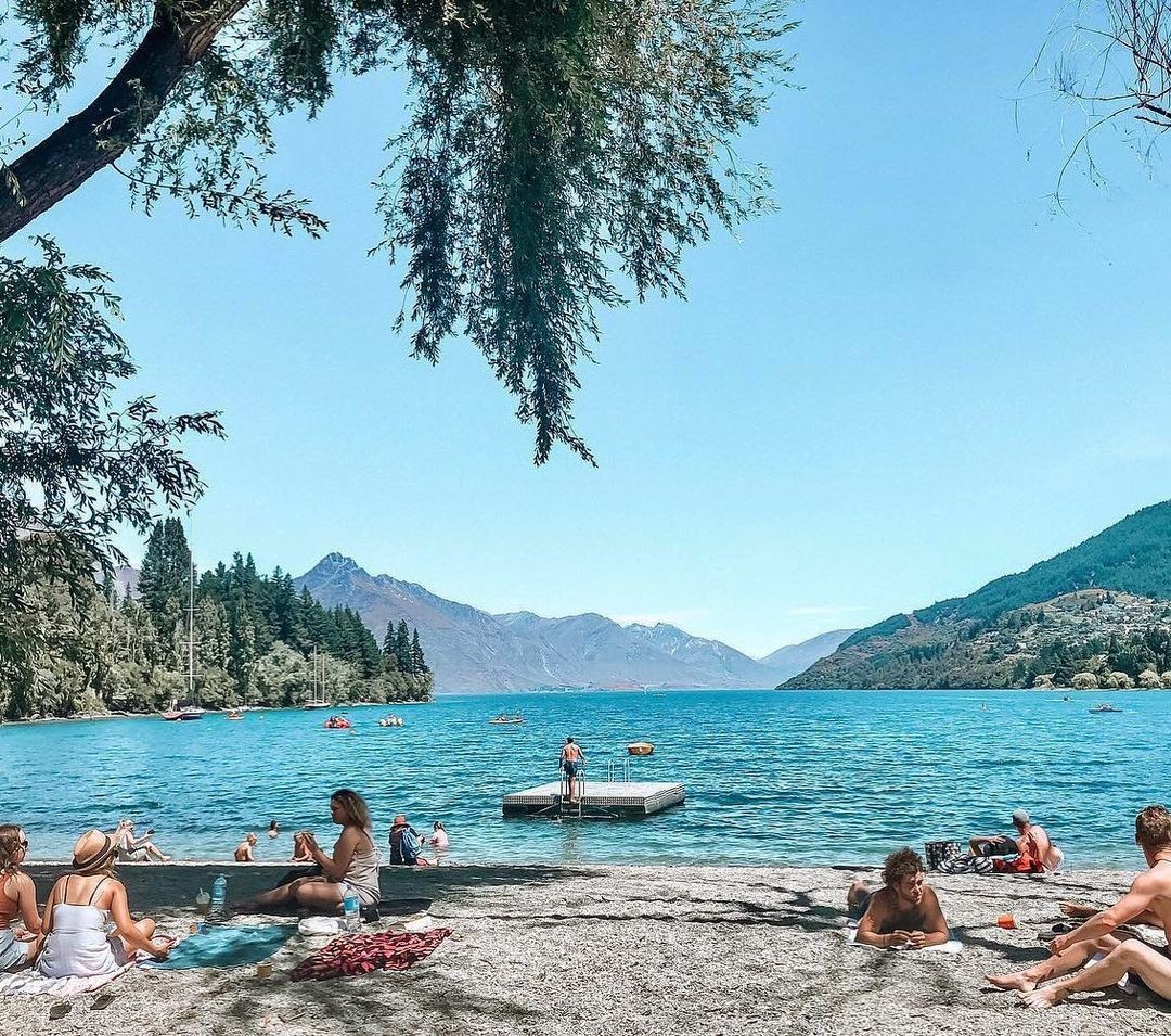 Groups of people sunbathe and swim at the Queenstown beach front during a summers day.