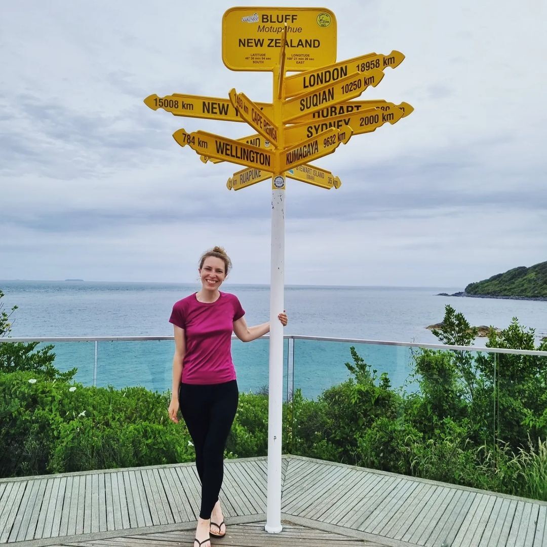 A woman stands next to the Bluff lookout point sign in Invercargill, New Zealand.