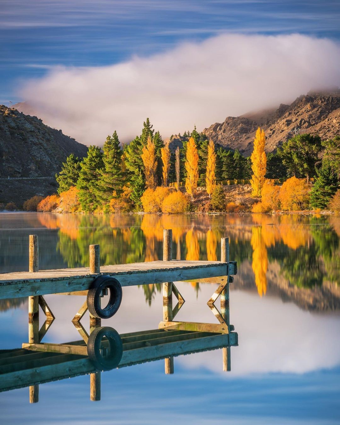 A stunning autumn capture of a wharf on Lake Dunstan with fall colours shimmering on the mirror reflection.
