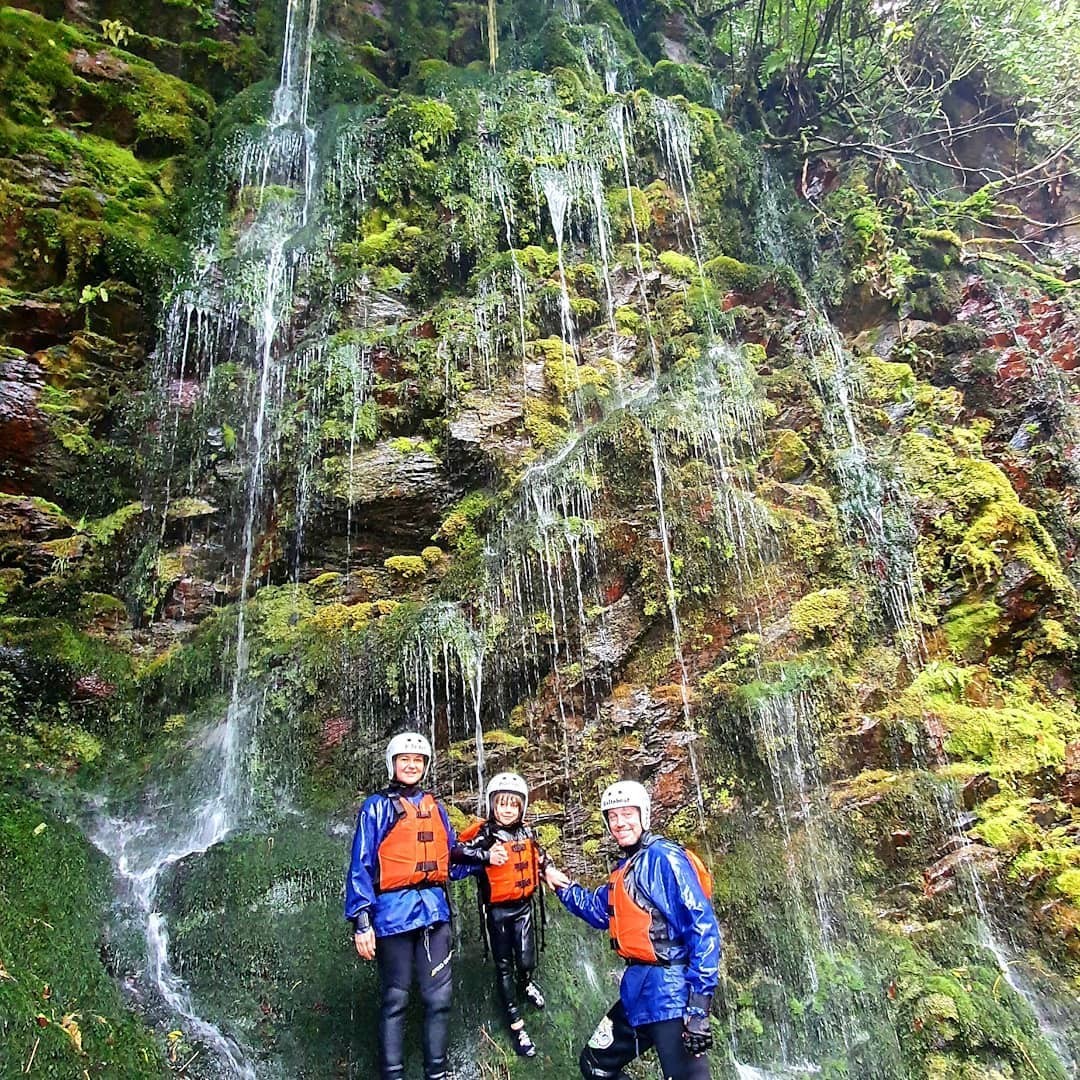 Three people stand beneath an impressive multi-tiered waterfall cascading down a moss-covered cliff side.