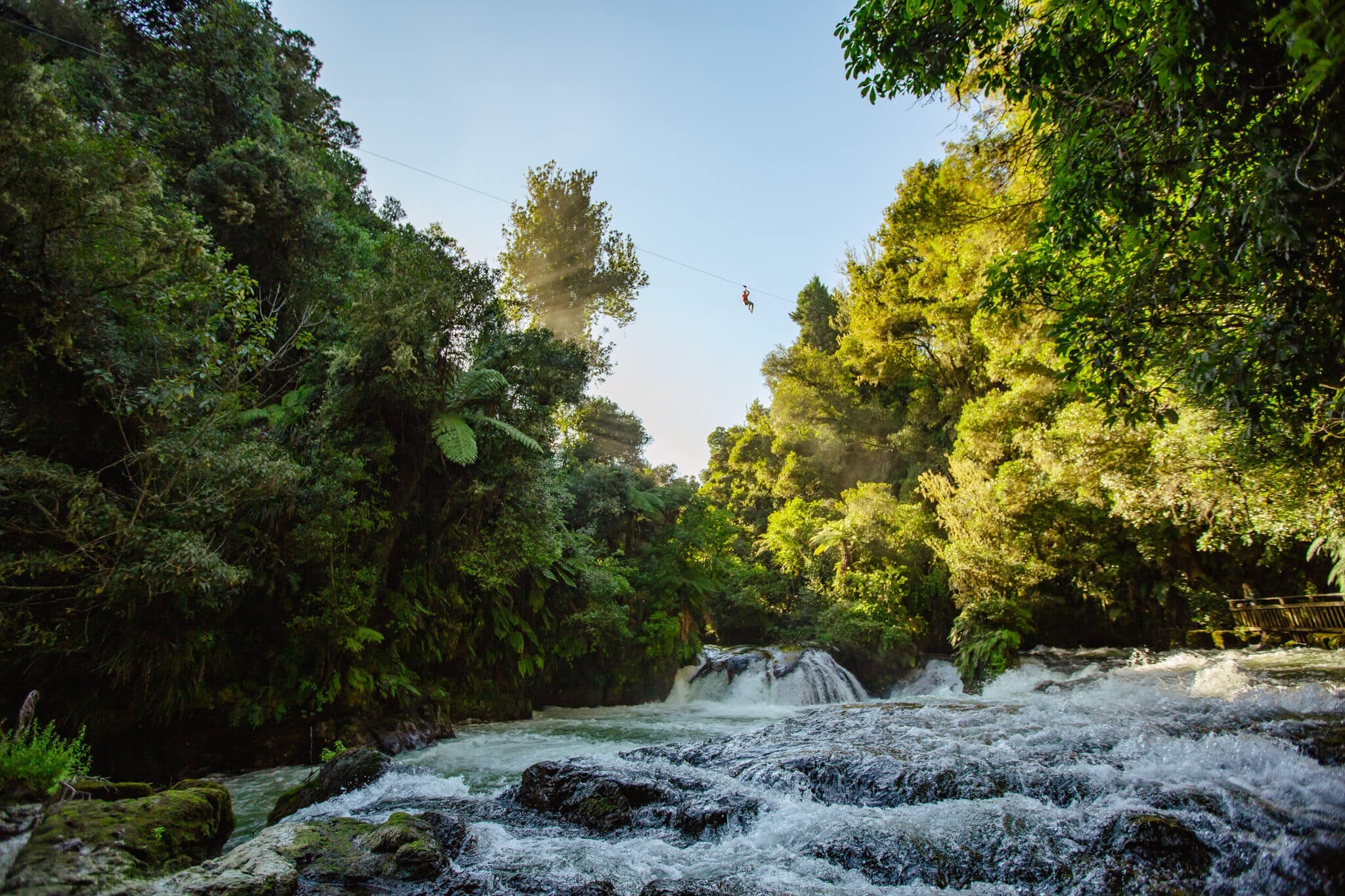 A person ziplines in the distance with the beautiful Ōkere Falls cascading in the foreground.