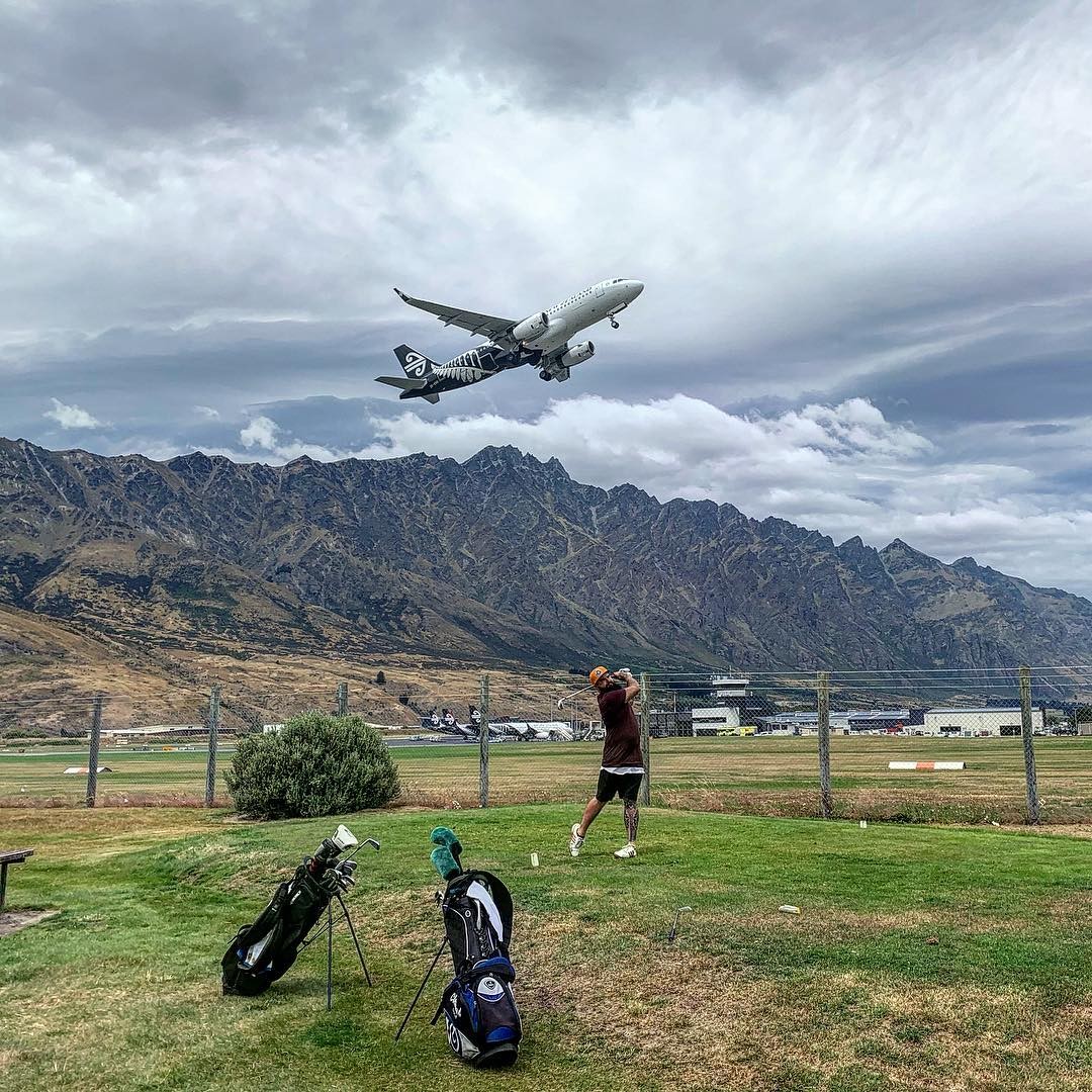 A man tees off at the Frankton Golf Centre with an airplane taking off in the back ground.