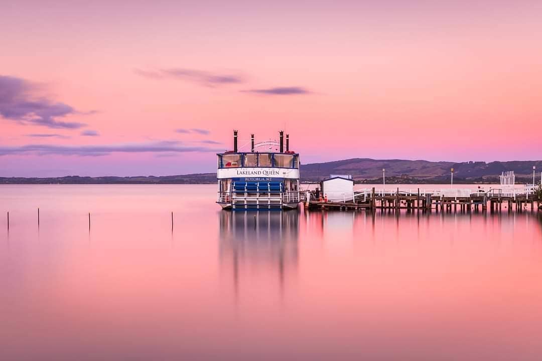 The Lakeland Queen parked at a wharf with a stunningly pink sunset on Lake Rotorua appearing behind.