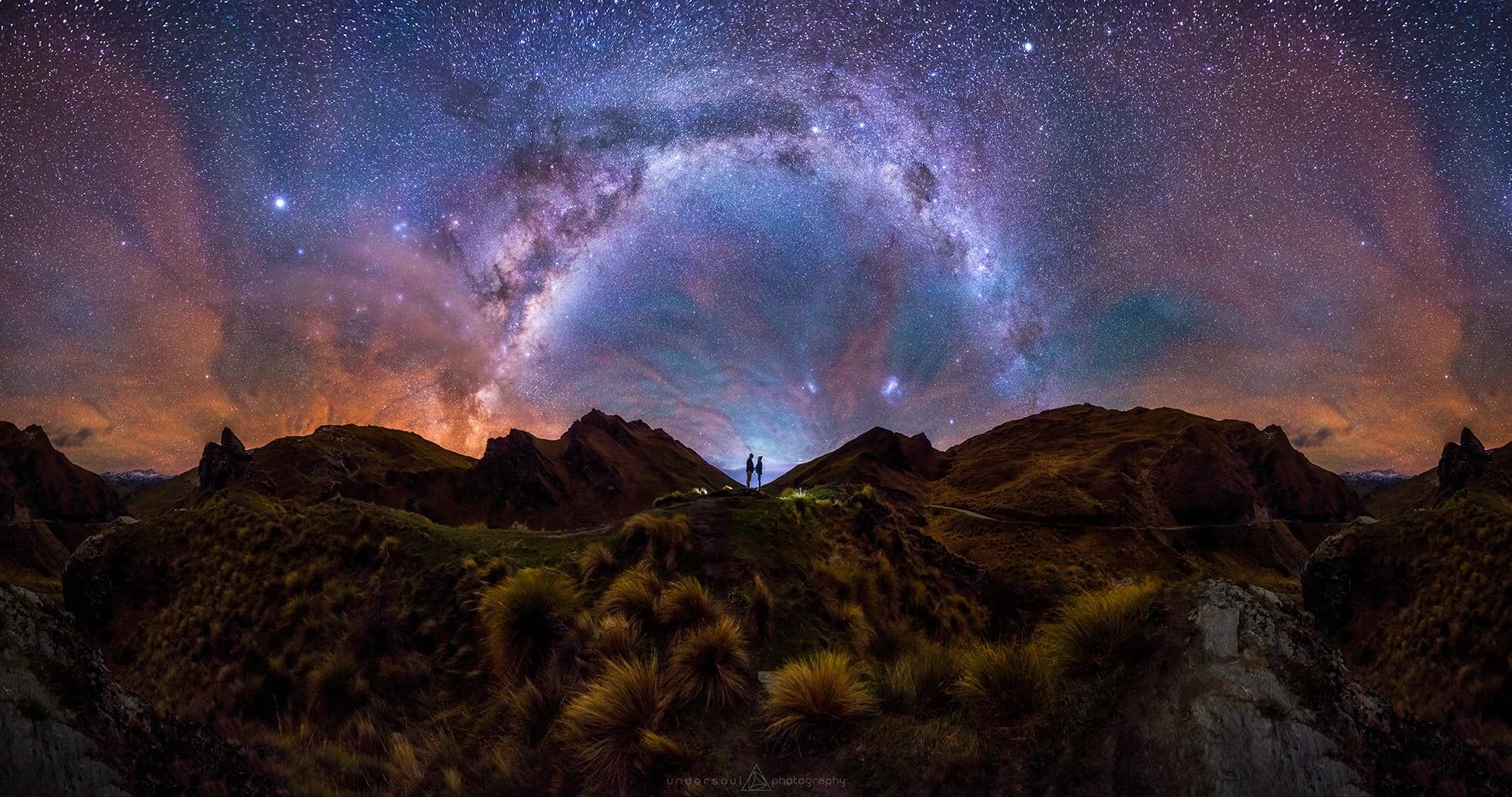 An incredible panoramic photo of the milkyway spanning the night sky with a couple standing in the middle of it.