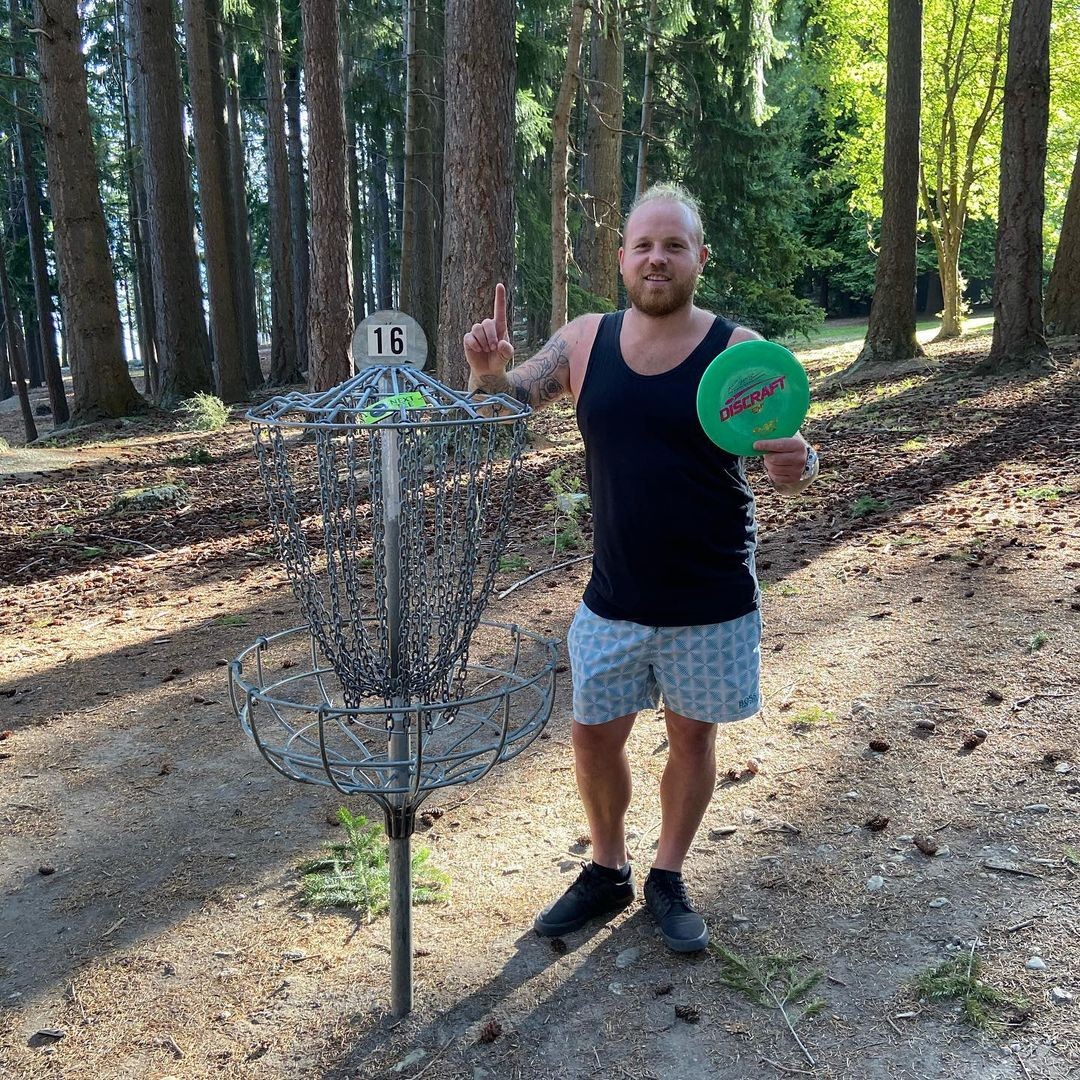 A man stands in front of a Disc Golf hole proudly holding his disc having completed the round.