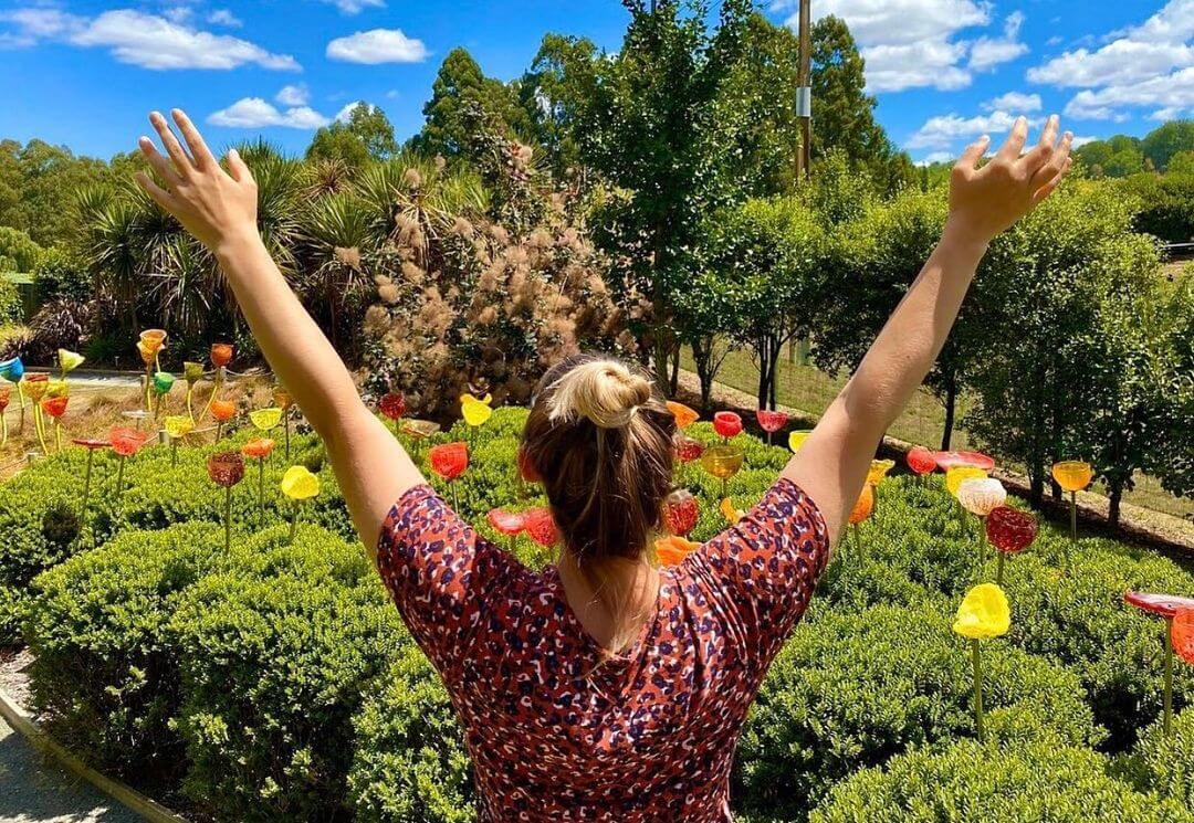 Woman stands in front of some flowers made from lava glass arms raised in the air.