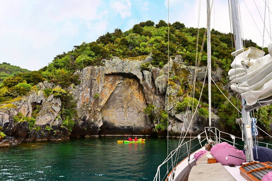 Family enjoy a kayak in front of the Maori rock carvings while a sailing boat waits for them.