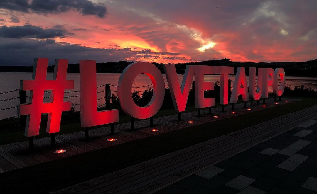 A lit up sign saying #lovetaupo with the sun setting in the background.