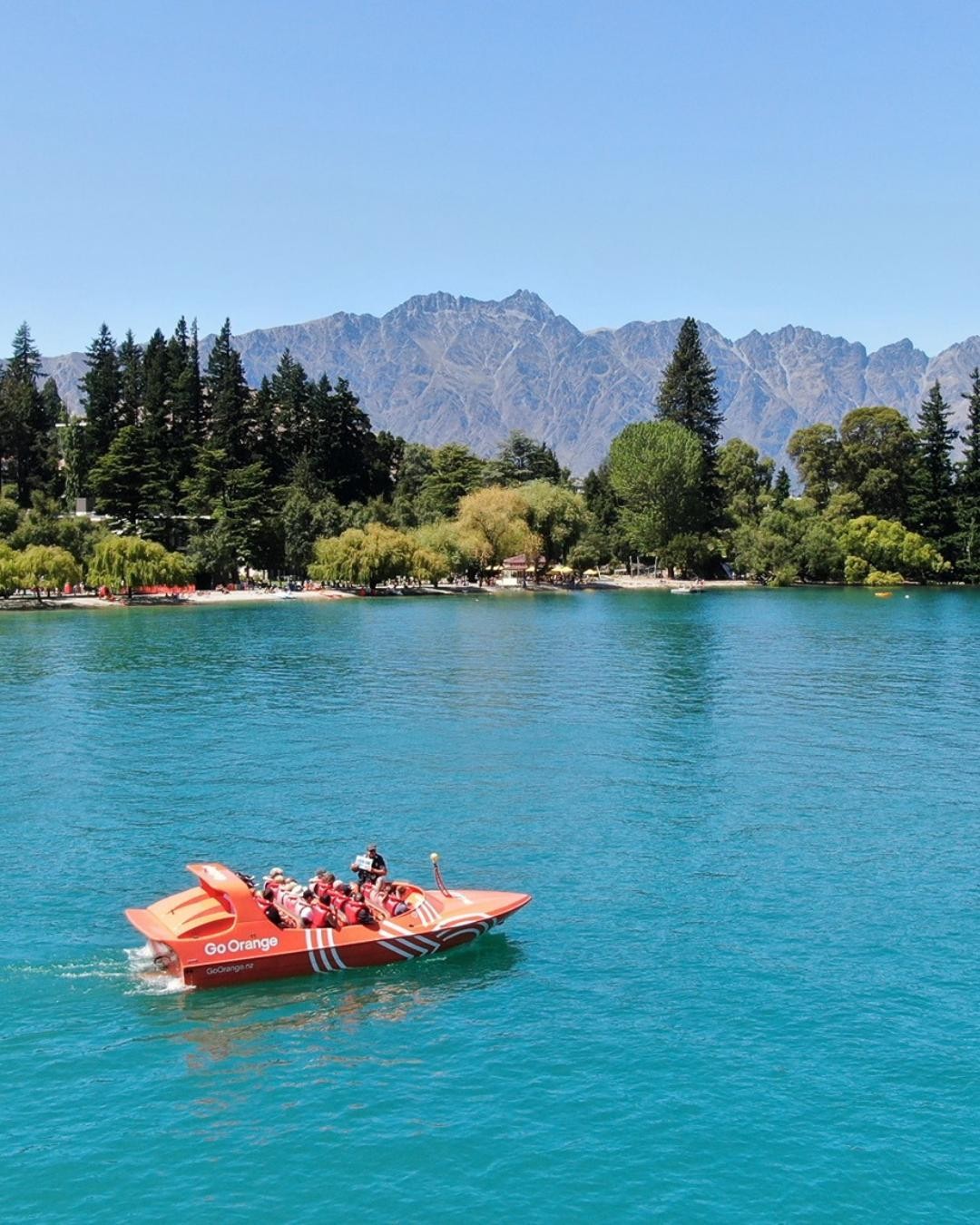 A jet boat cruises Lake Whakatipu in summer with views of The Remarkables mountain range behind.