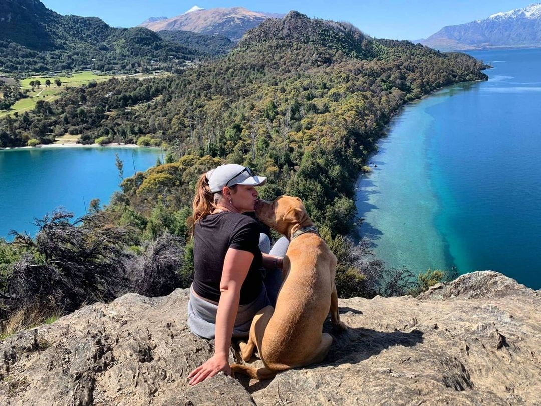 A lady kisses her dog with beautiful view of Bob's Cove, Queenstown behind them.
