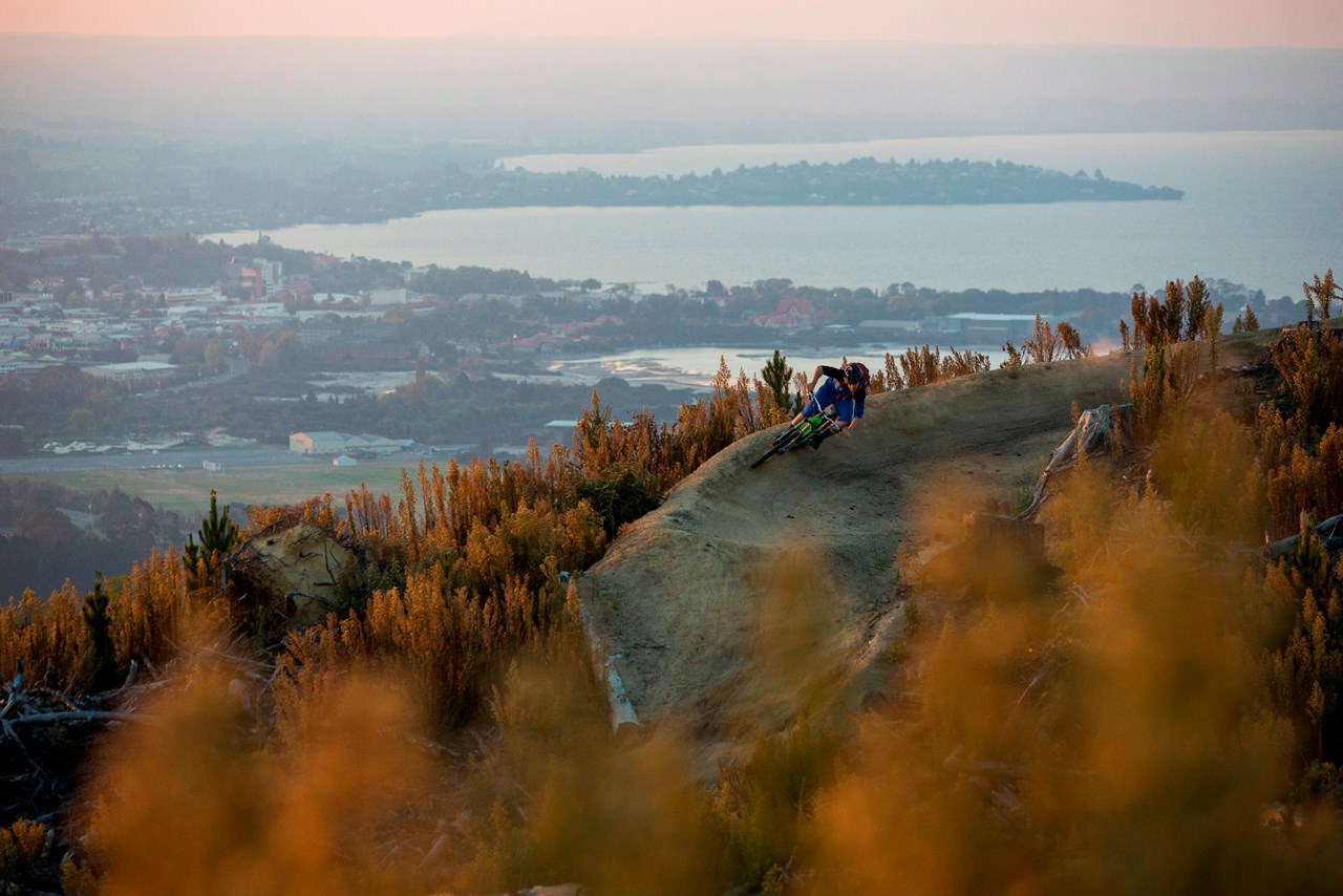 A mountain biker navigates a mountaintop corner with panoramic views of Rotorua behind them.