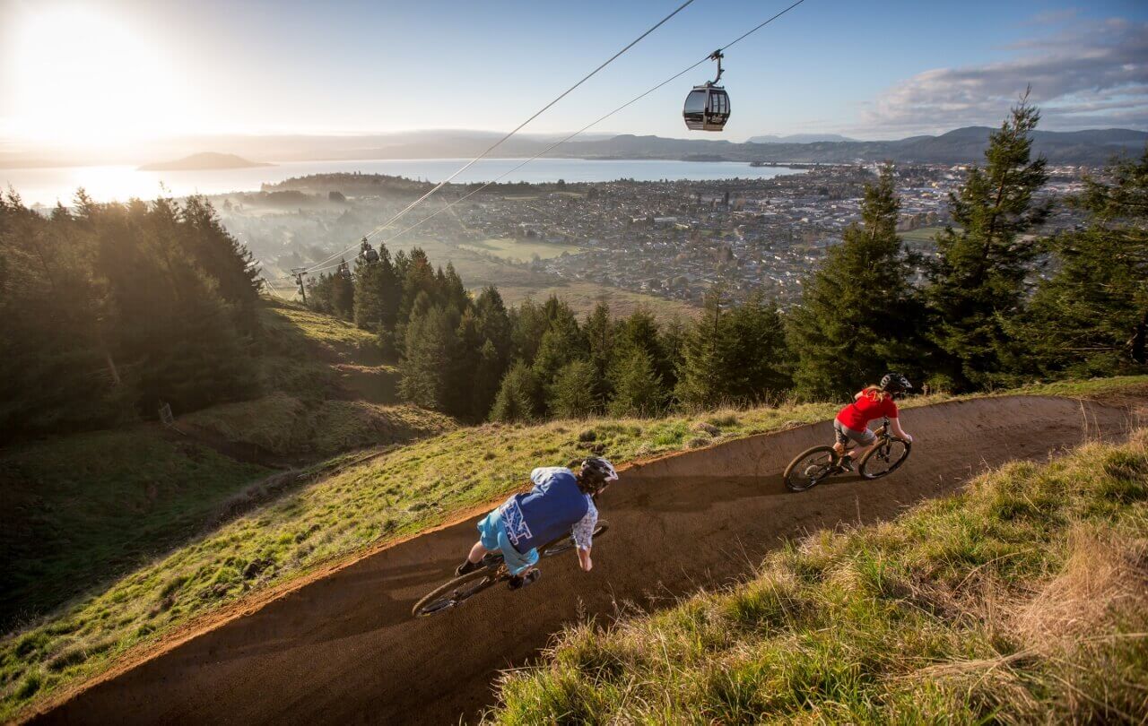 Two bikers riding riding on the gravity park overlooking Rotorua and the Skyline Gondola on a sunny day.