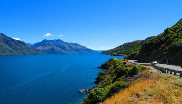 The road from Kingston to Queenstown stretches along with the Lake on one side and greenery on the other, all under a blue sky.