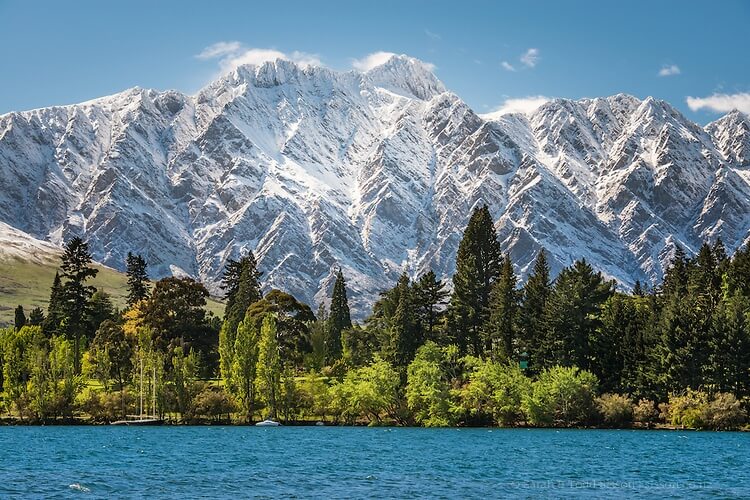 A snow covered Remarkables mountain range sit behind green gardens and trees, and a blue Lake Wakatipu.