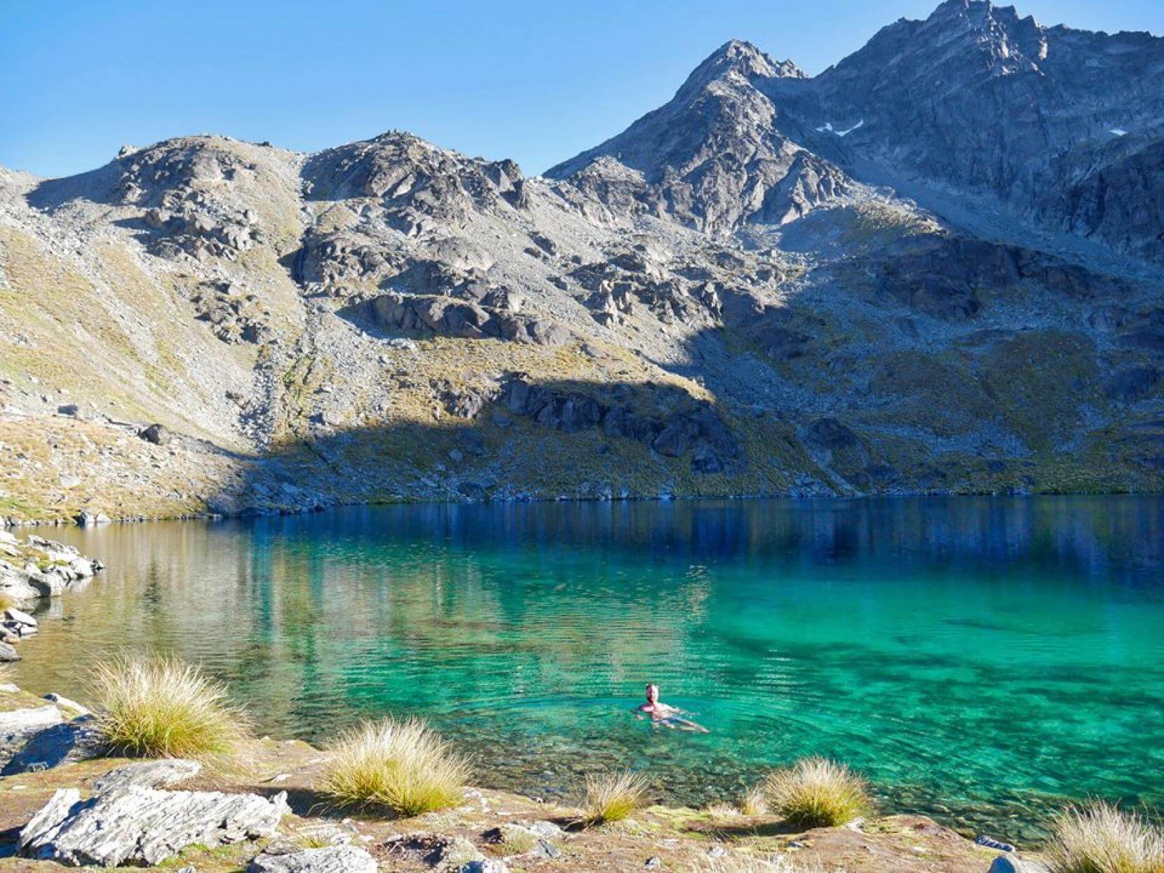 A man swims in Lake Alta at Remarkables, with mountains in the background.