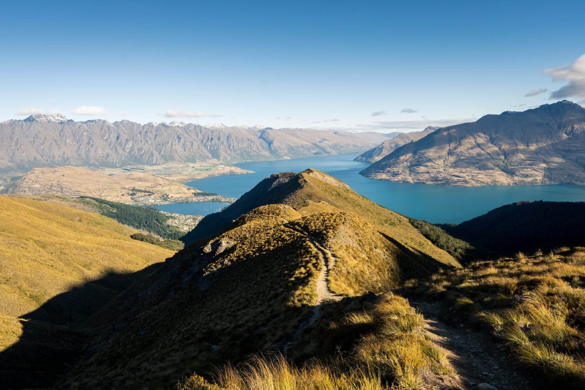 A walking track leads along the peak of a long mountain, with views of the surrounding mountains and lake.
