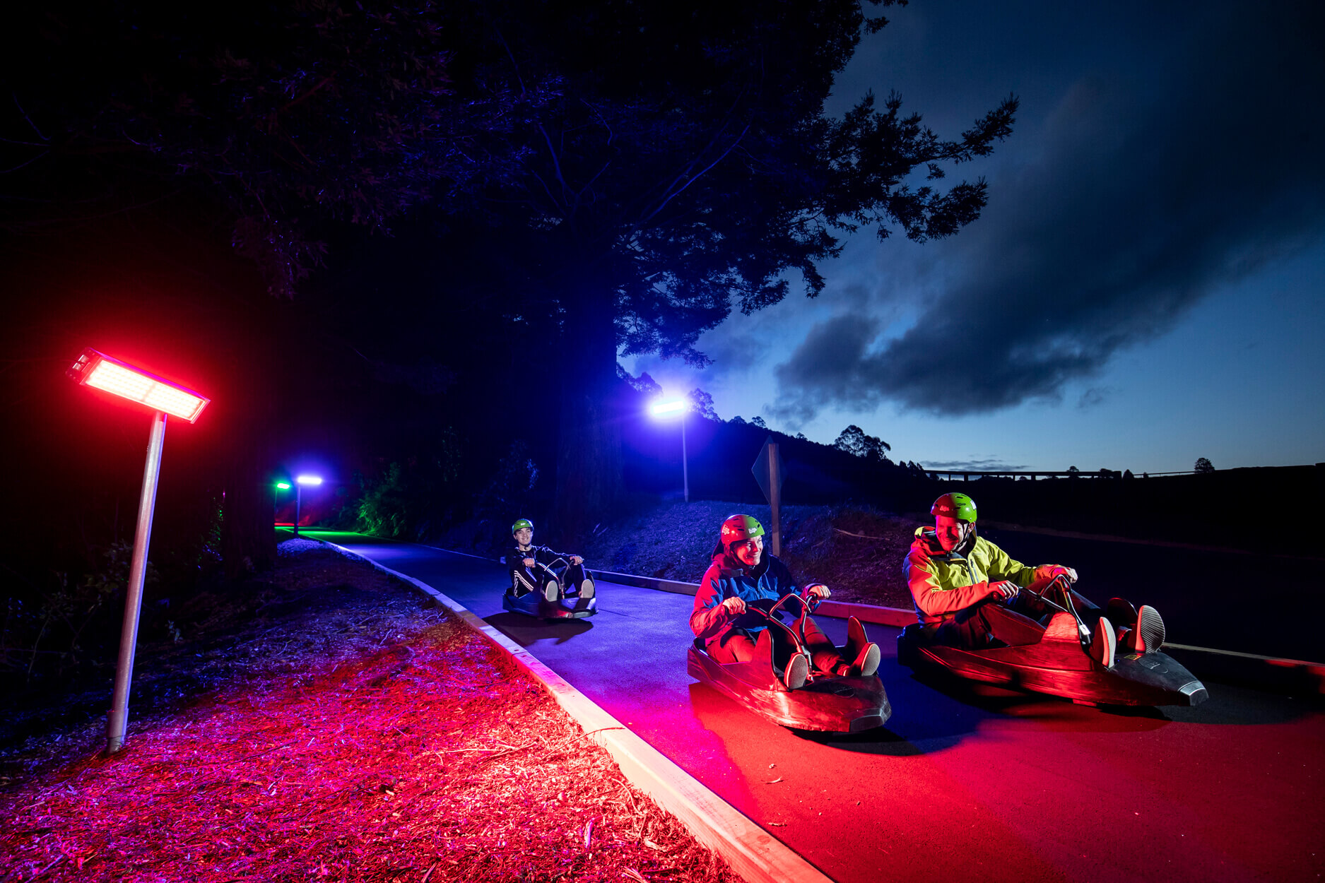 Three riders luge down the track at night, illuminated by red and blue lights.