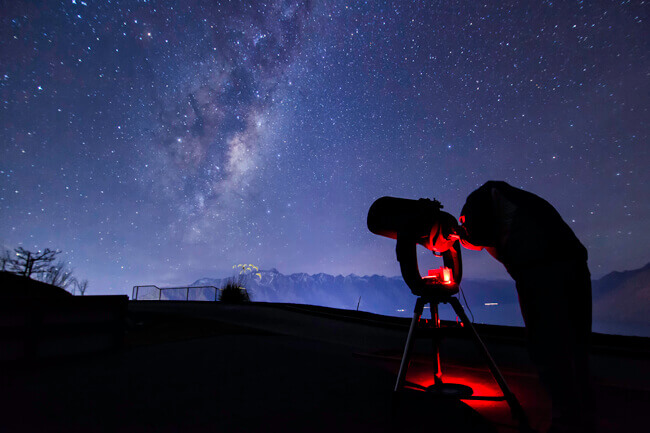 A person looks through a telescope at night at Skyline Queenstown while gazing upon the Milky Way which is lighting up the sky above them.