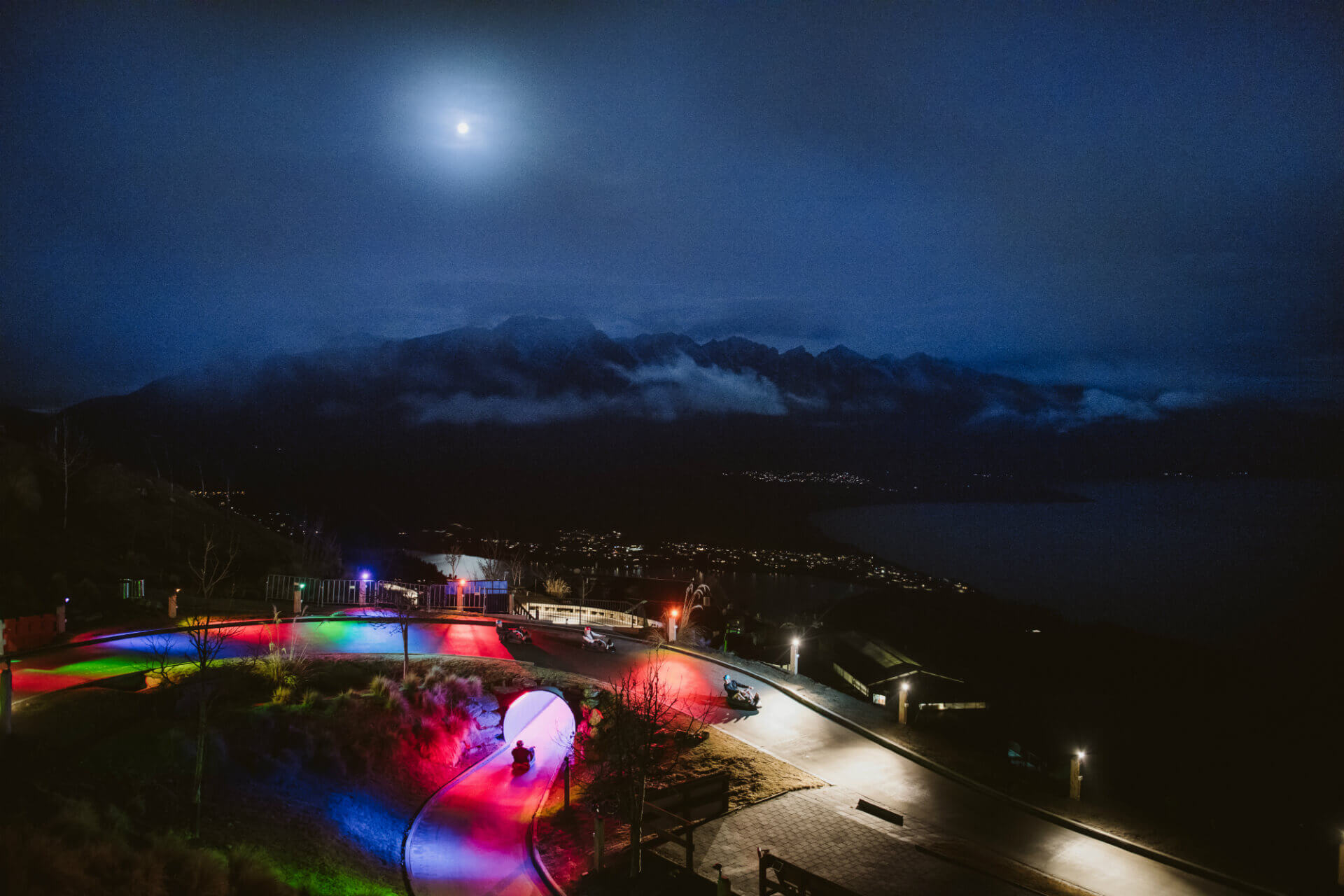 Section of Luge track lit up by colourful lights at night, with the moon high above lighting up the surrounding mountains.