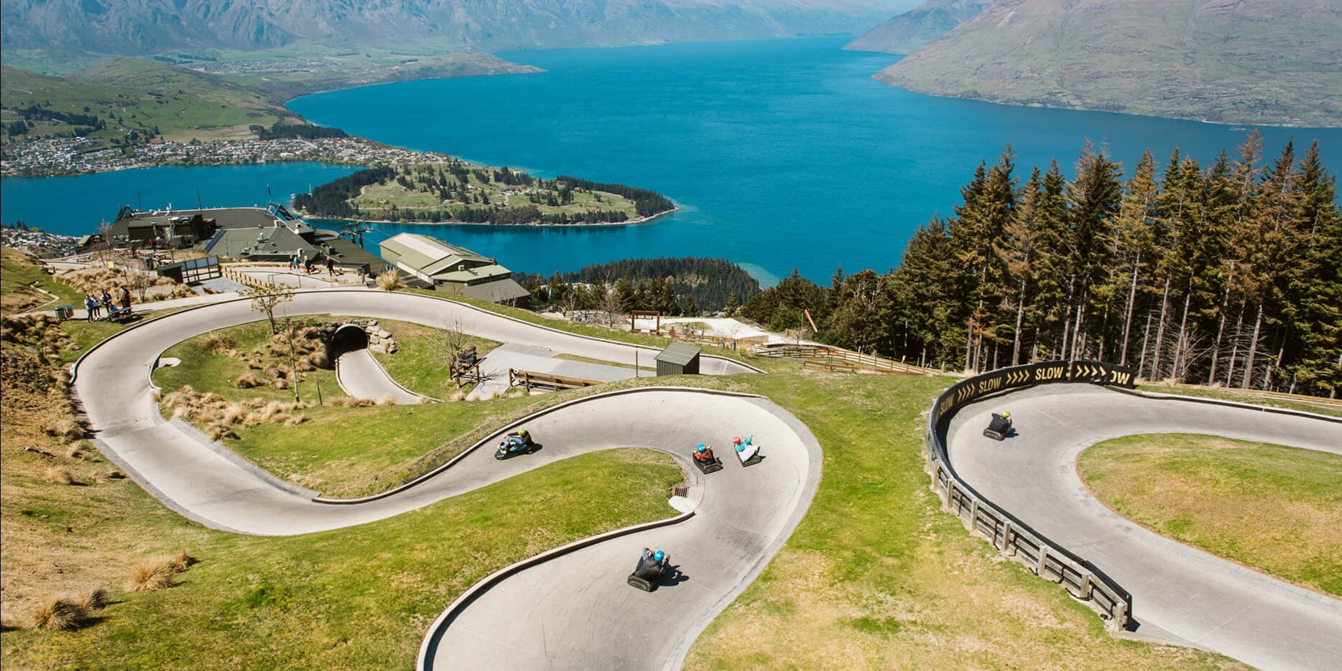 Aerial view of luge riders racing down twisted path at Skyline Queenstown with Lake Wakatipu and Queenstown Gardens visible in the distance.