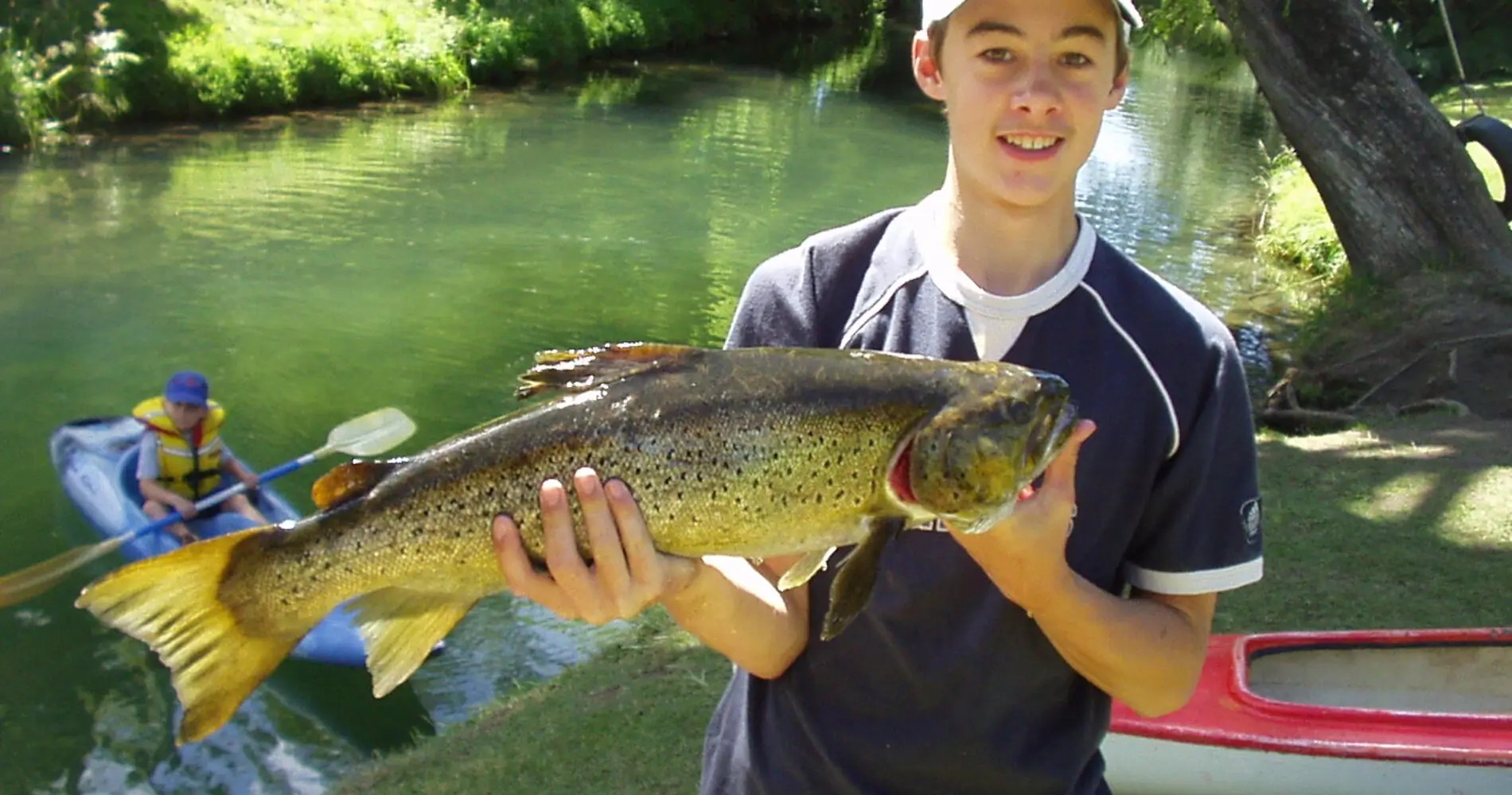 Young angler proudly displaying large trout catch beside stream with kayakers and boat in background at holiday park.