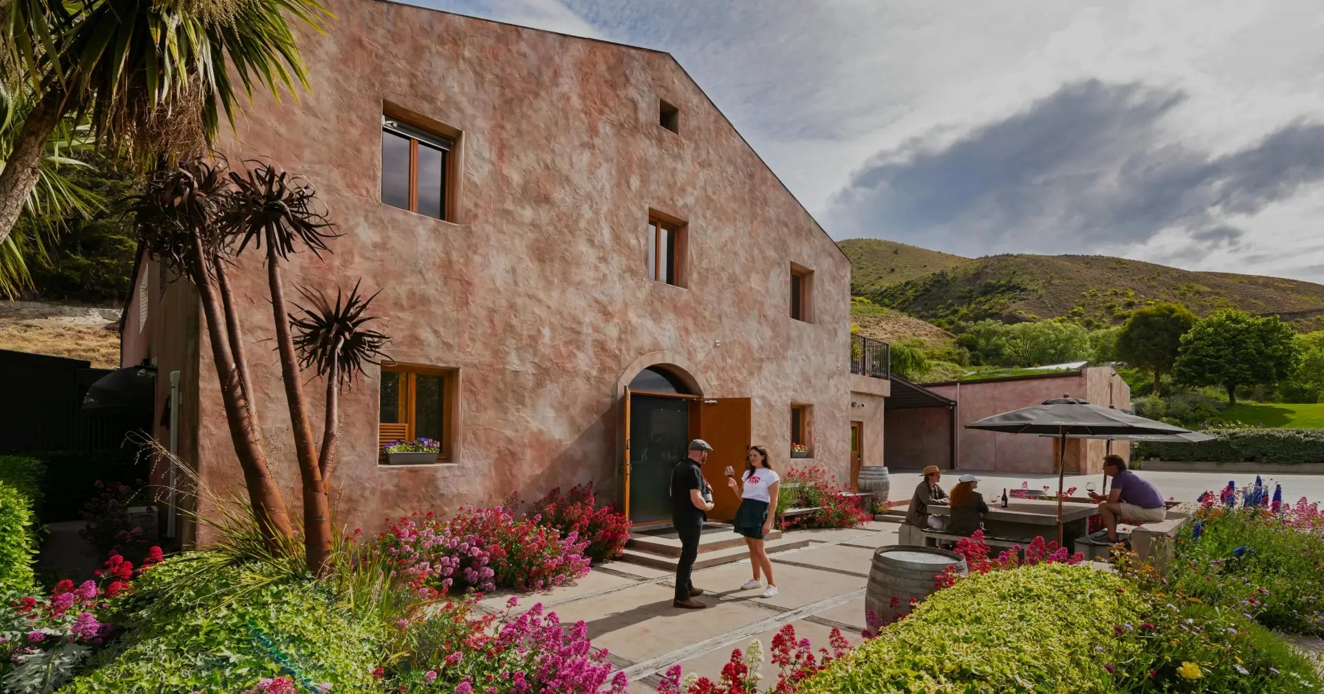 Rustic stone winery building with landscaped gardens, palm trees, and visitors exploring the scenic vineyard grounds.
