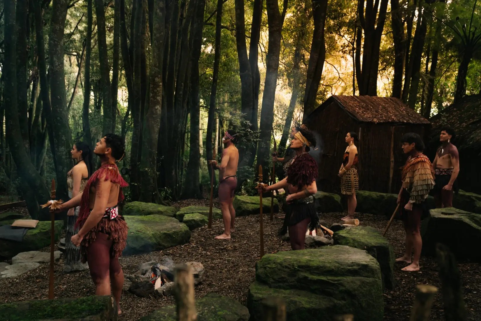 A group of Māori performers standing in a forest clearing and holding taiaha spears for the Summer Harvest Celebration at Te Pa Tu, Rotorua.
