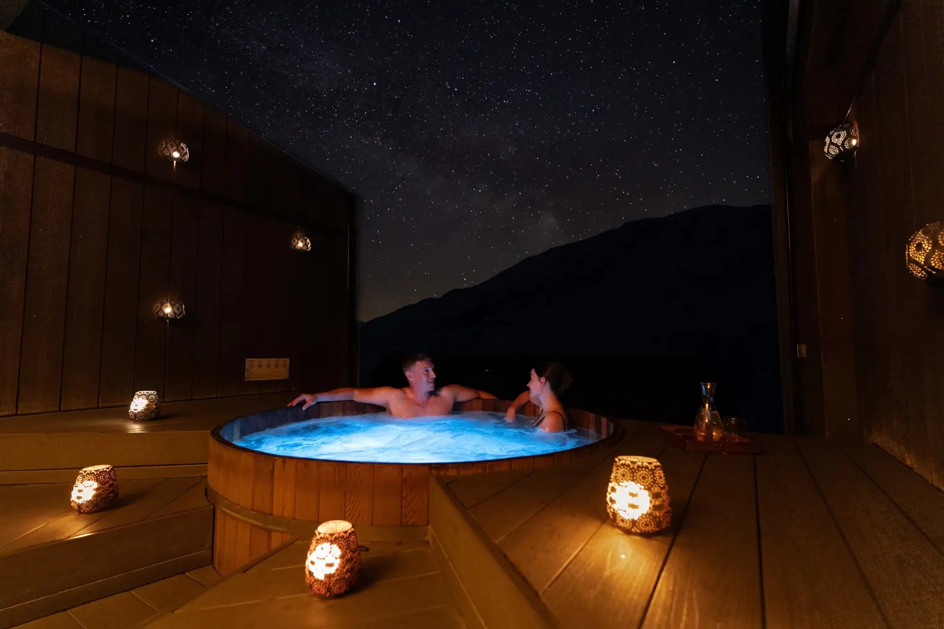 A couple in a lit-up spa pool in the open air, surrounded by lantern lights at Onsen Hot Pools in Queenstown.