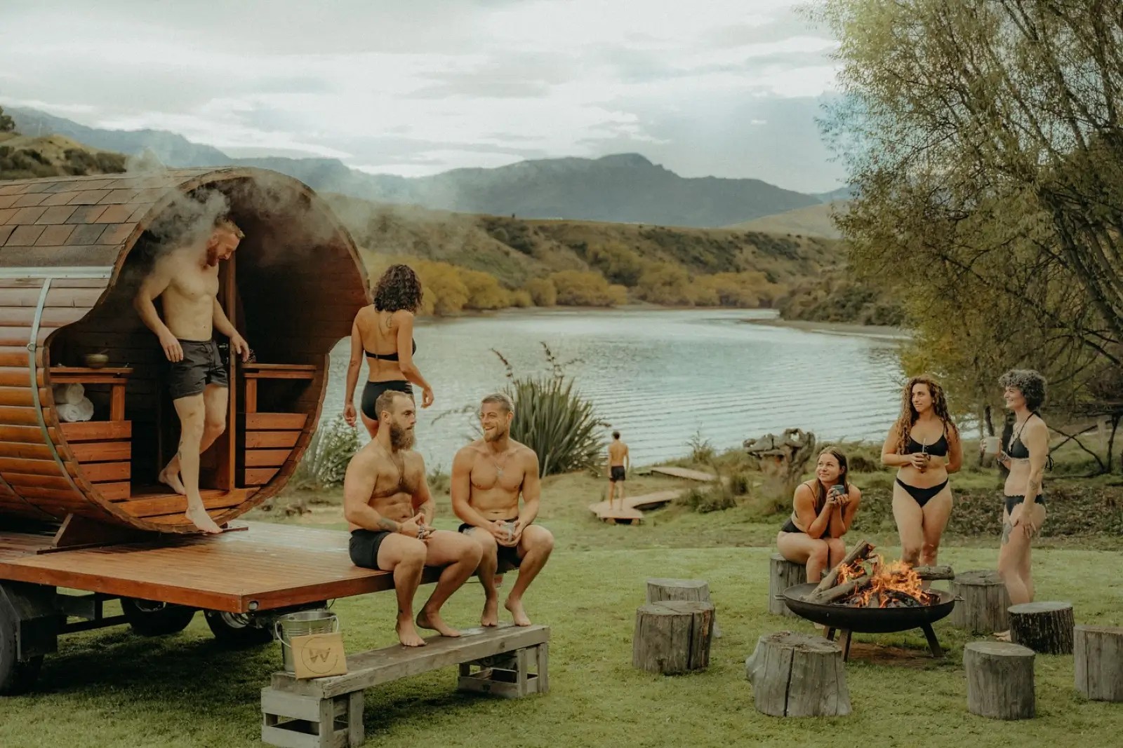 Two people exiting a wooden portable sauna, two people sitting on the edge of the sauna's deck, and three people holding mugs next to a fire nearby.