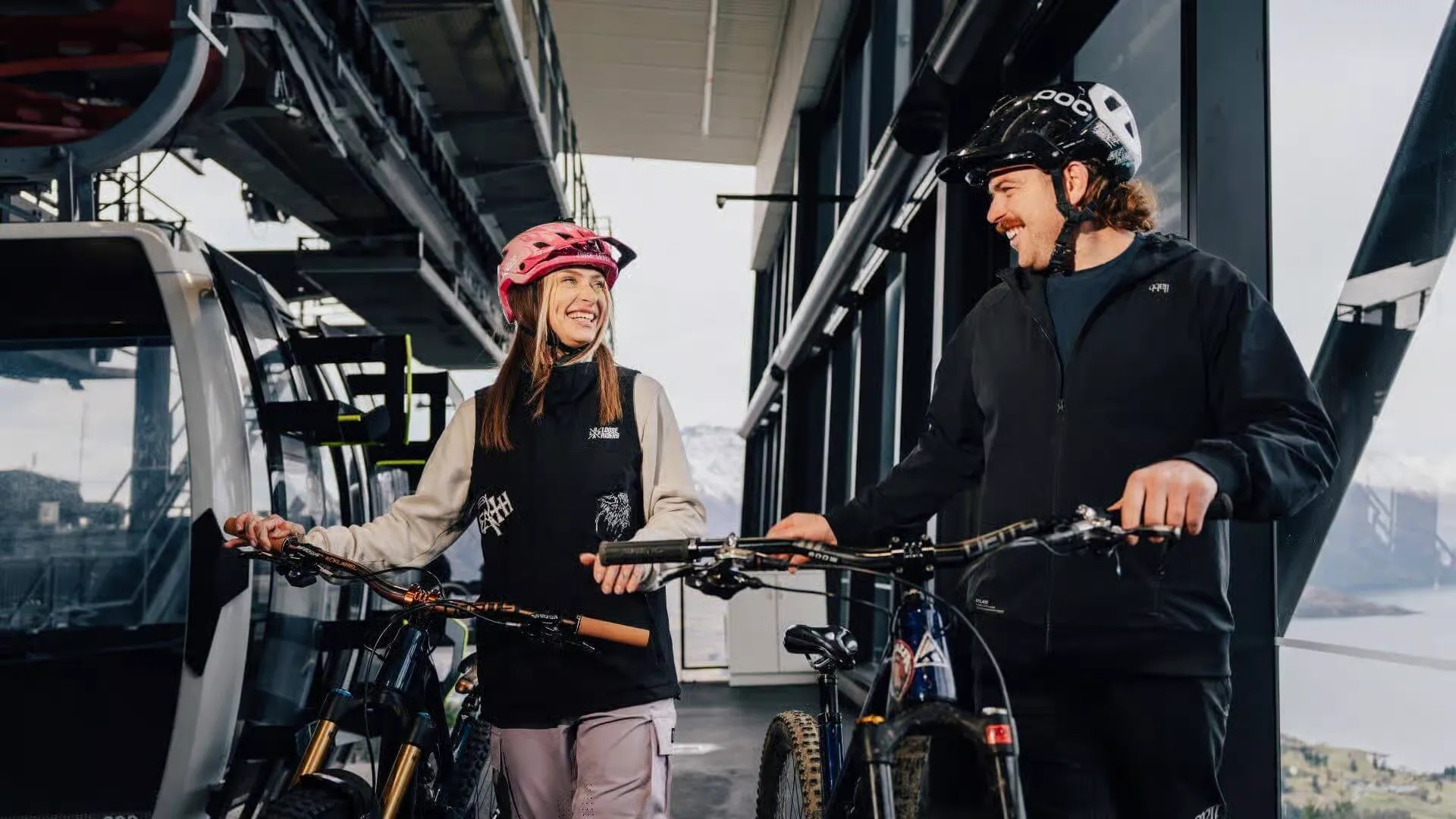 Man and woman smiling at each other at the Skyline Queenstown Mountain Bike Uplift gondola platform. Each individual is wearing a helmet and guiding a bike in their hands.