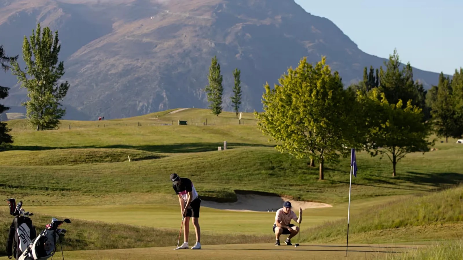 In an expansive green golf course with a mountain in the distance, a person putts near a hole while another watches.