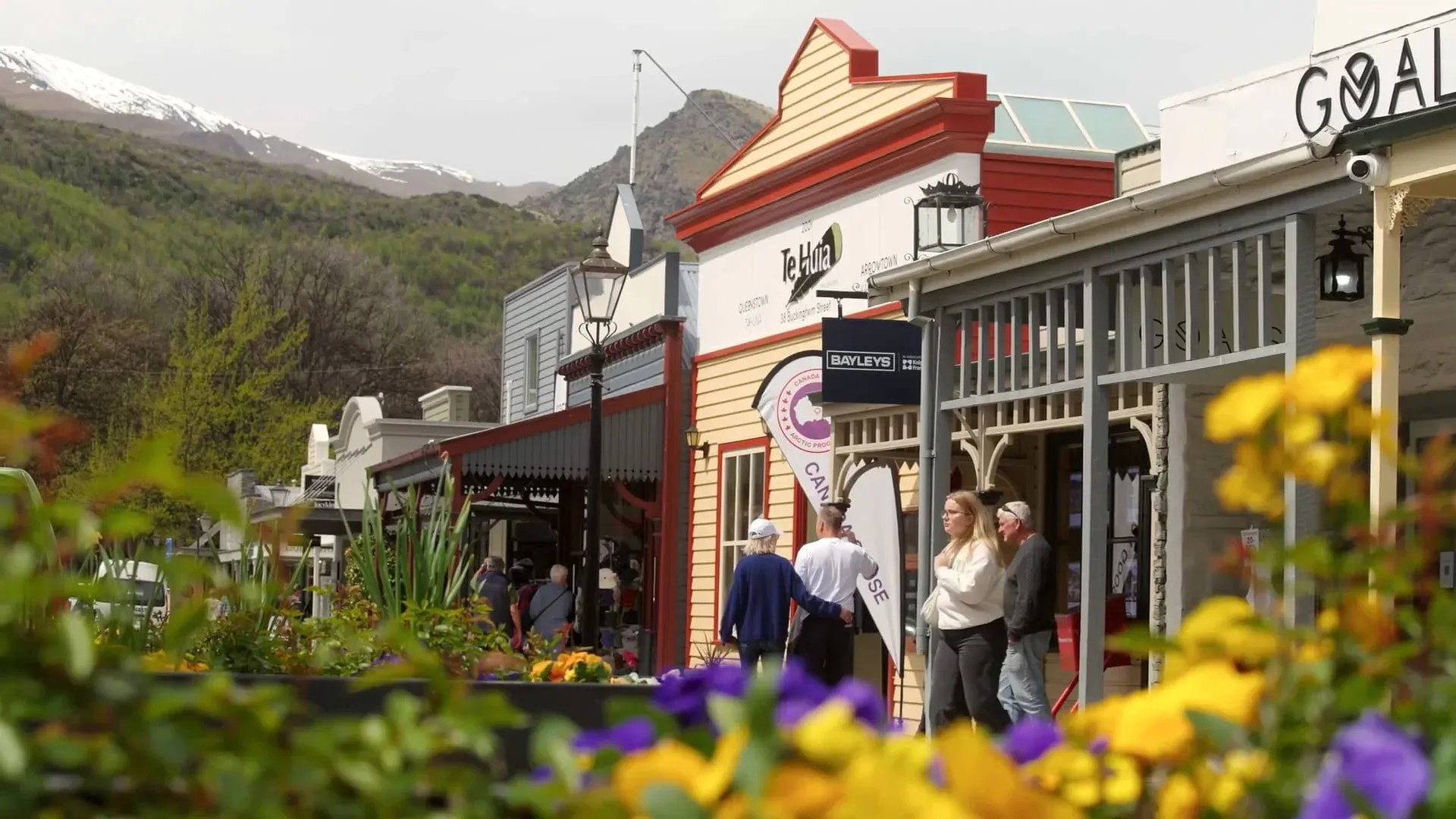 Street in Arrowtown with flowerbeds, shops with historical architecture, and pedestrians walking around.