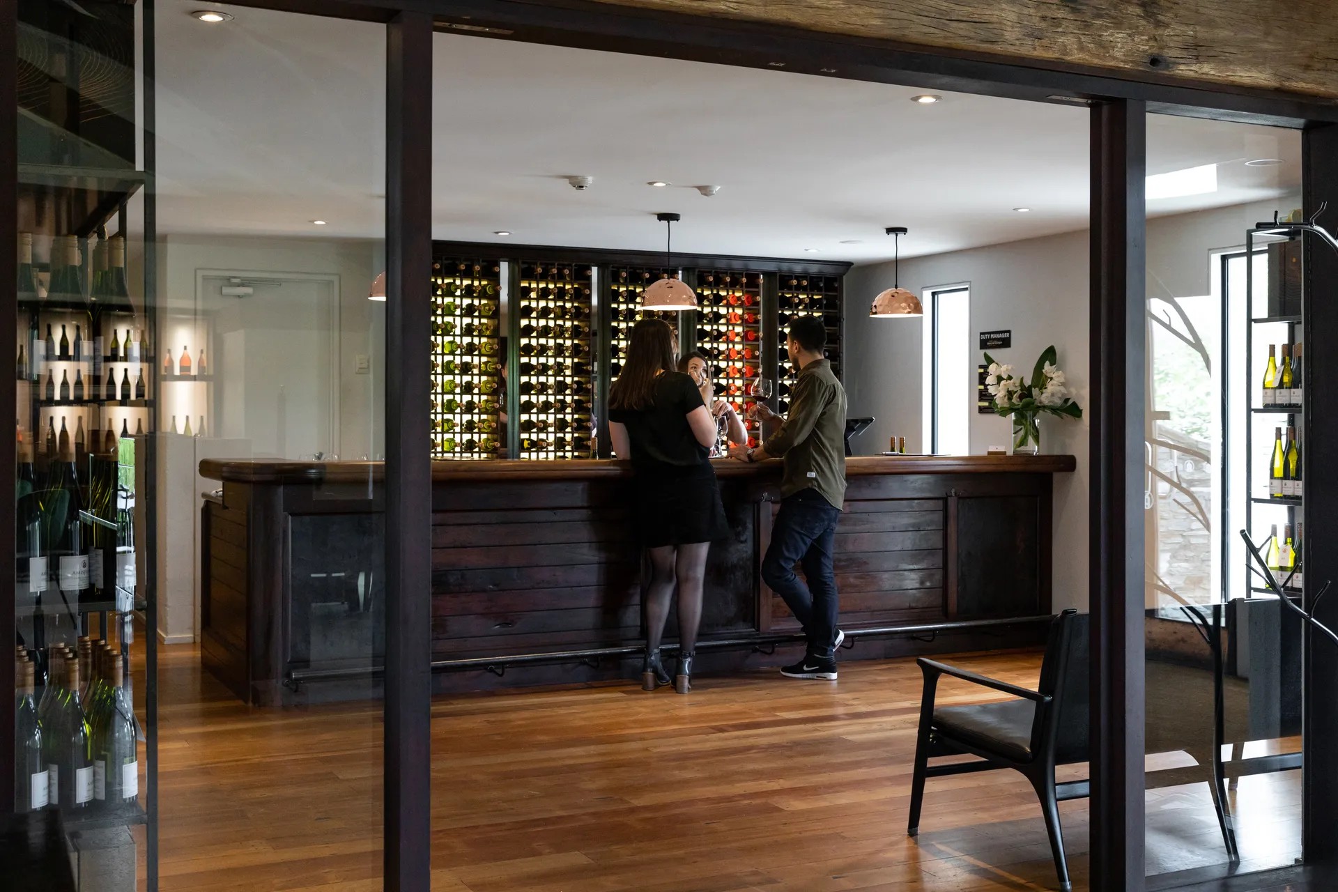 Stylish winery bar area with guests sampling wine, extensive bottle display wall and warm timber furnishings.