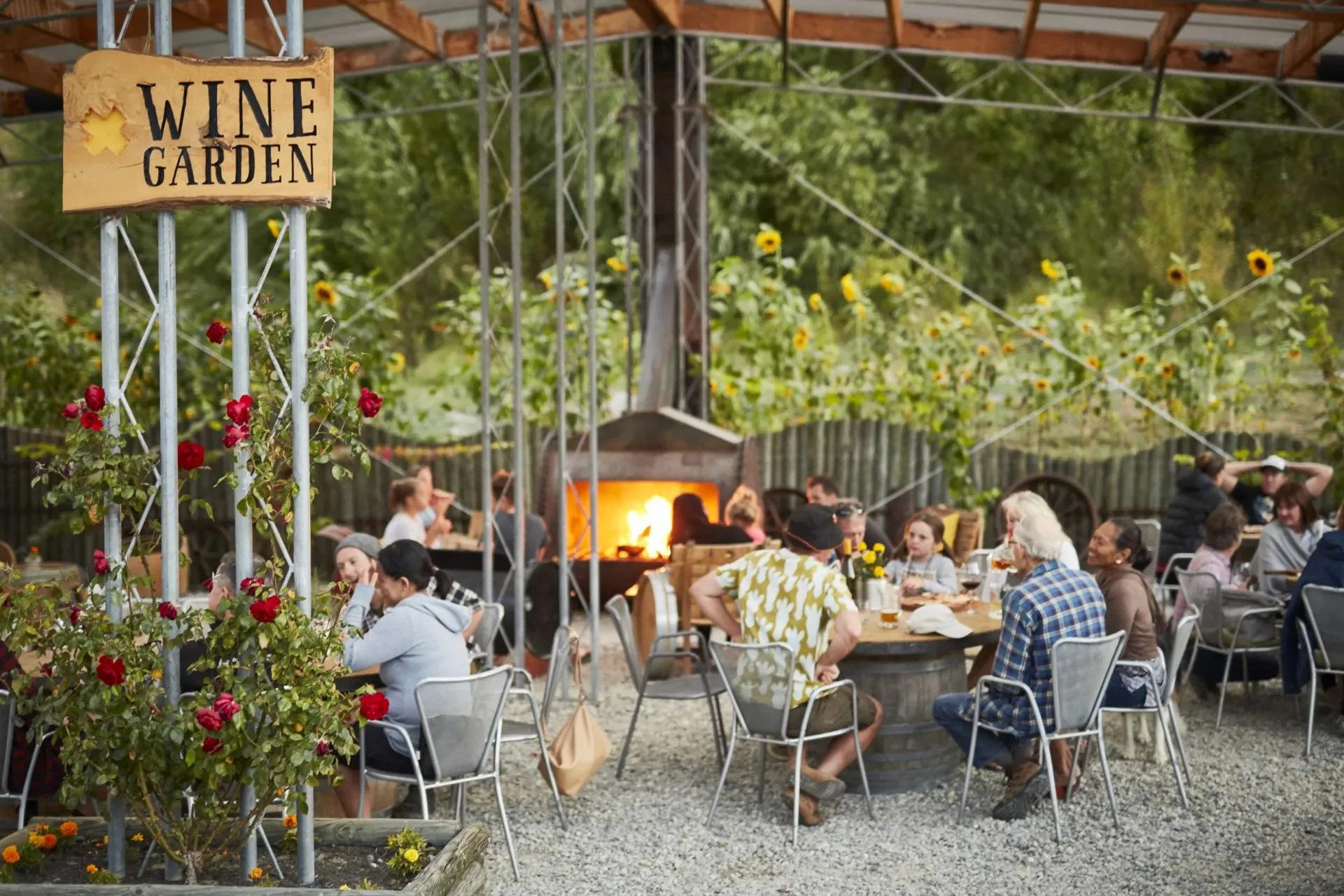 Guests drinking wine in a rustic wine garden with barrel tables and flowers.